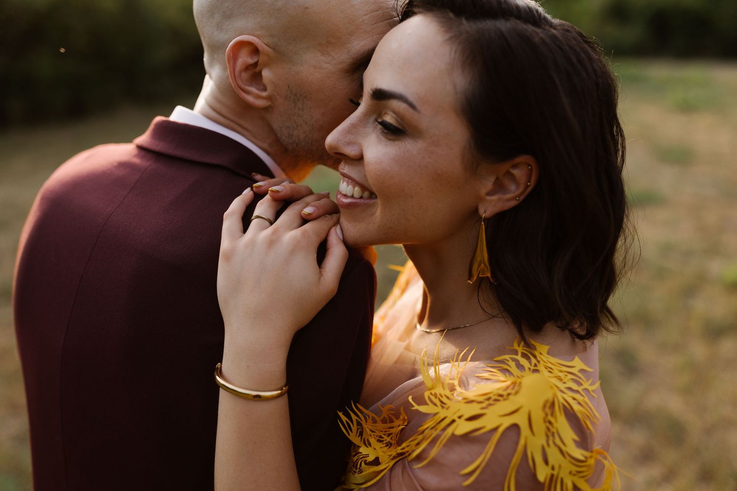 Couple sharing an intimate moment in a yellow dress and burgundy suit against a natural backdrop.