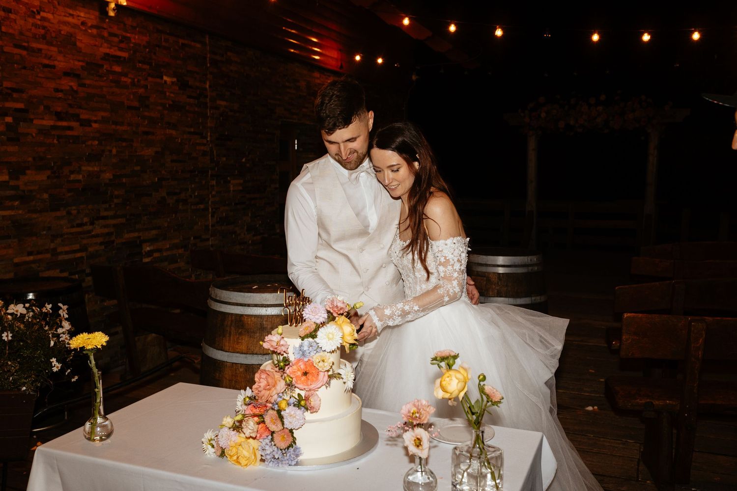A couple shares an intimate moment while cutting their floral-decorated wedding cake at a rustic venue with string lights.