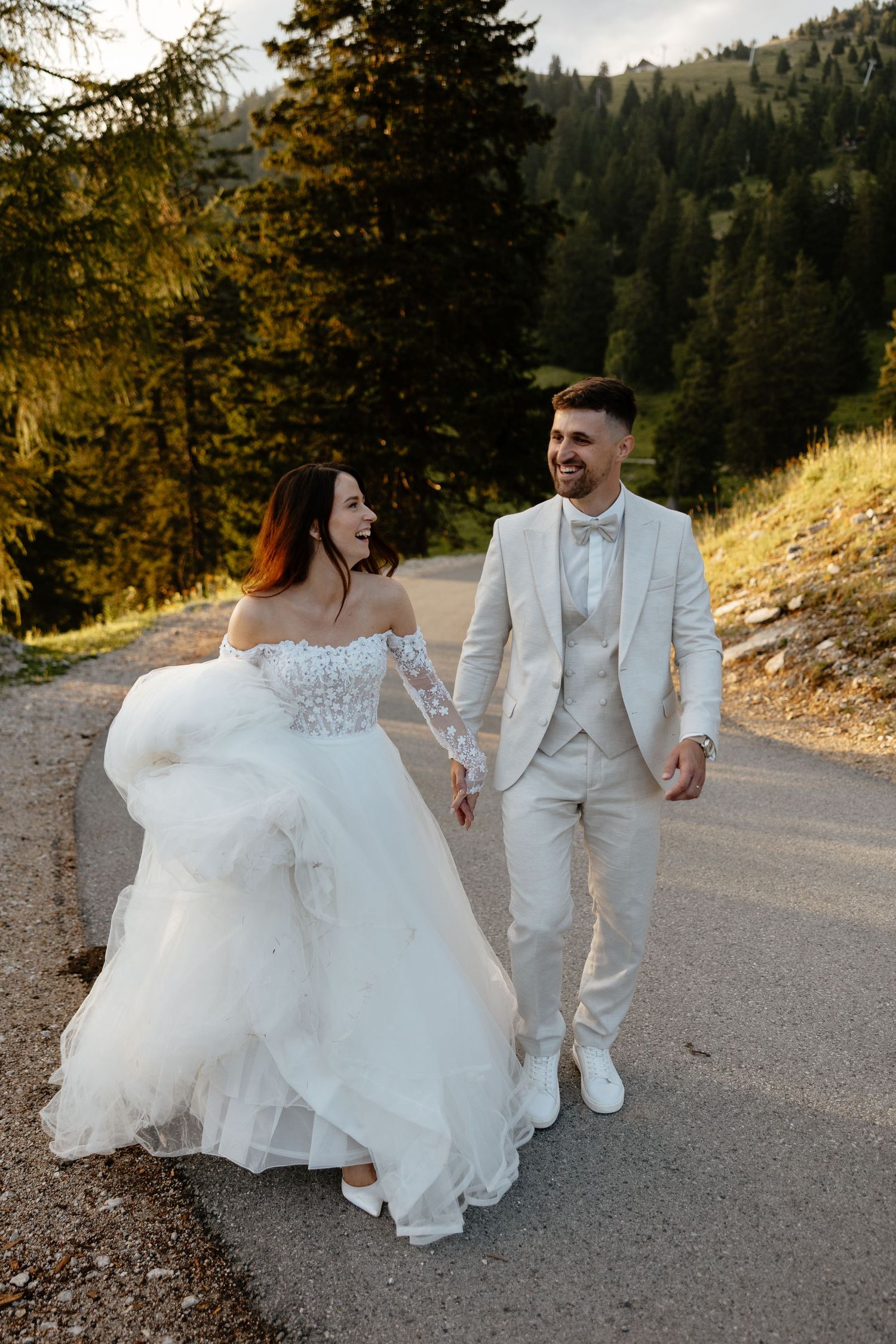 Newlyweds walking hand in hand on mountain path with bride's dress flowing in the breeze.