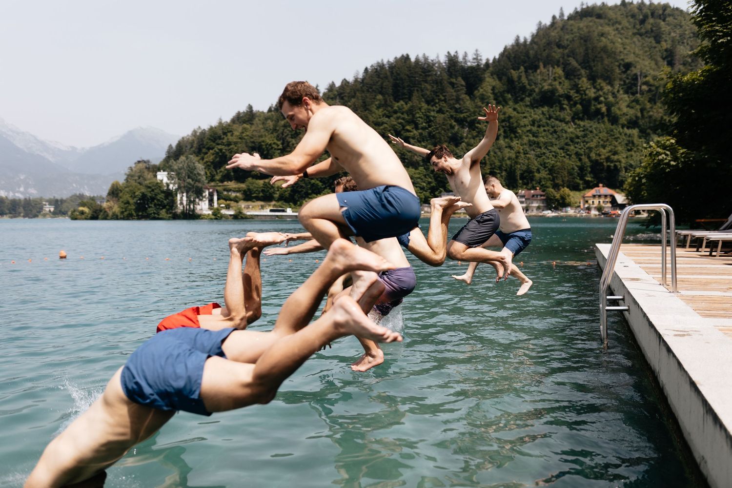 Group of friends jumping into a scenic mountain lake from a wooden dock on a summer day.