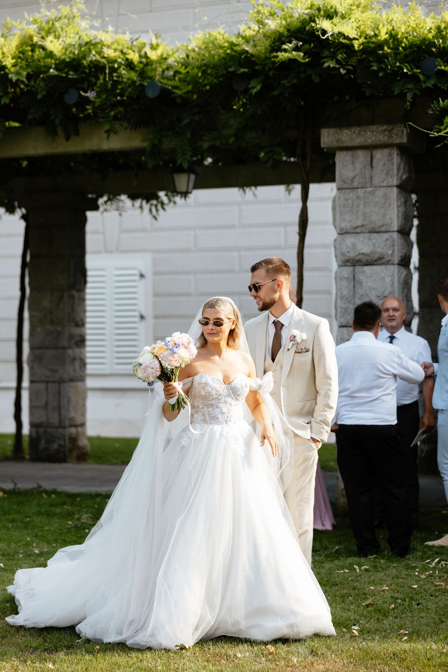 Wedding party poses for photos outdoors near white buildings with beautiful floral bouquets.