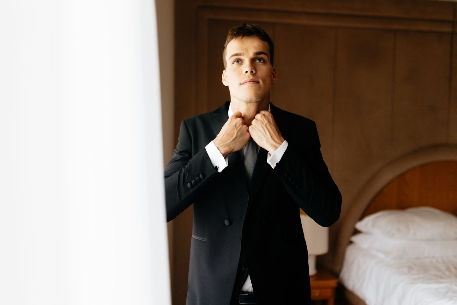 A person in a dark suit sits thoughtfully in a dimly lit room with their hand near their face.