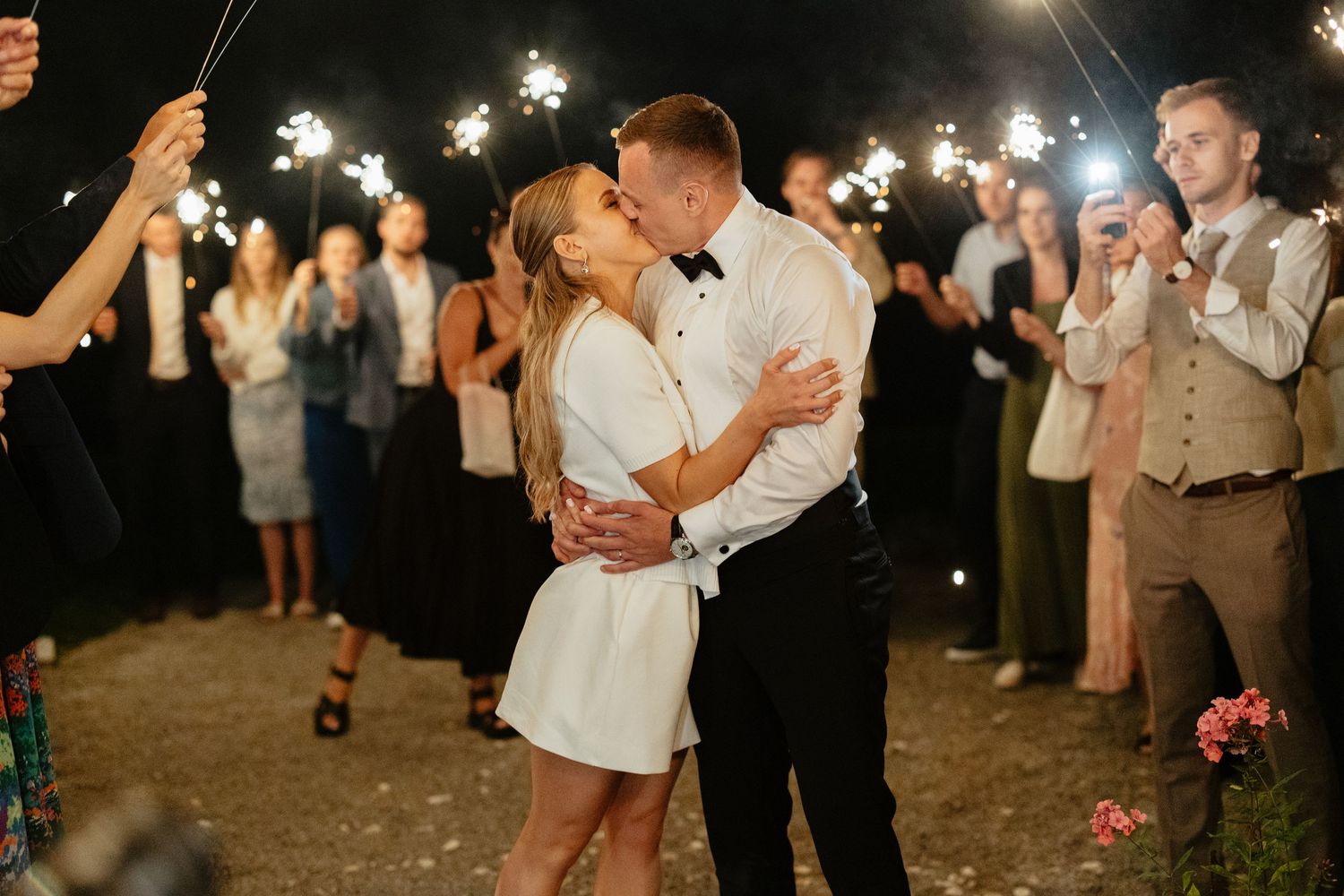 Newlyweds share a romantic kiss surrounded by sparklers held by wedding guests at night.