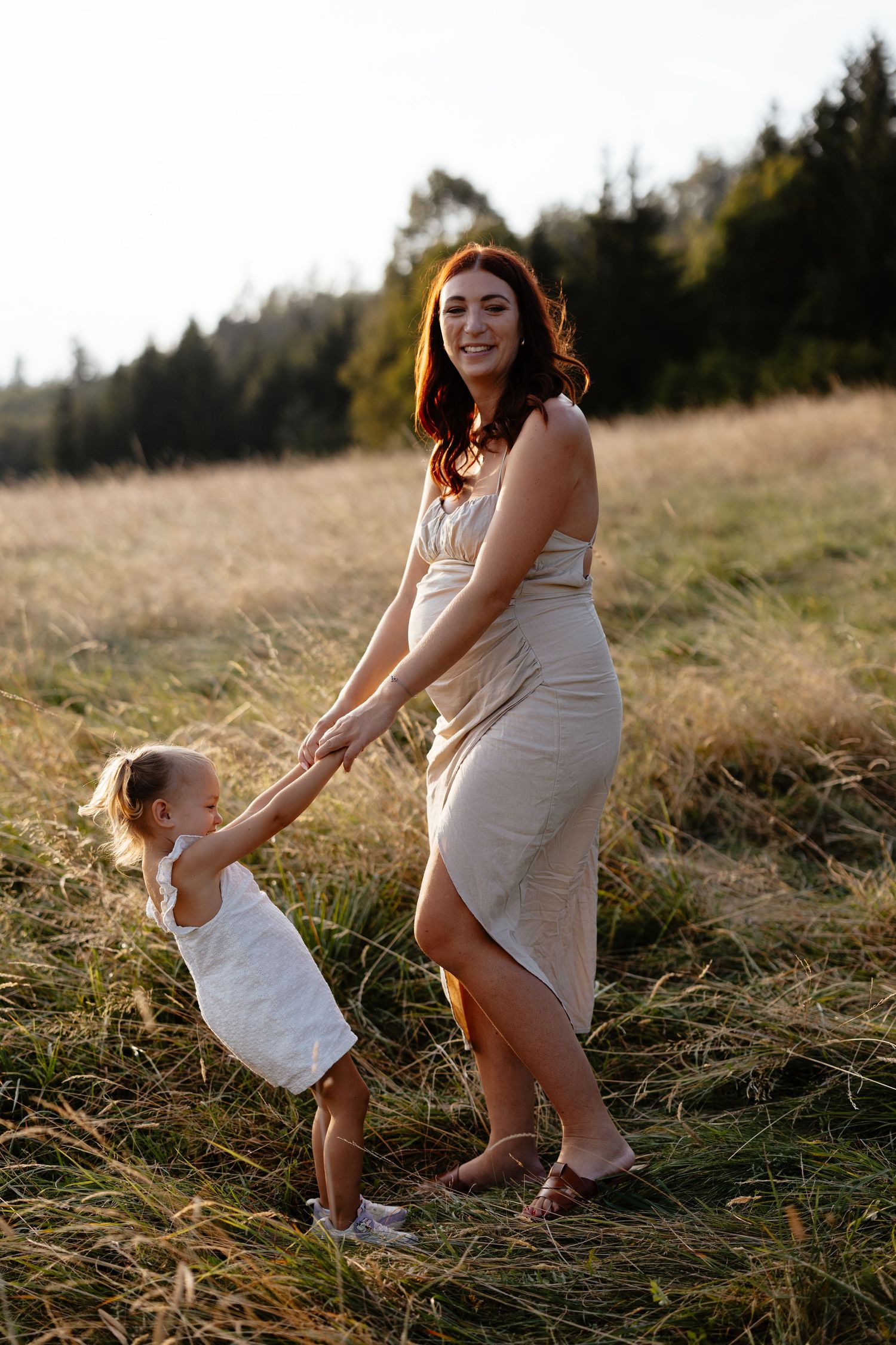 A mother and child in white dresses dance together in a sunlit field at golden hour.