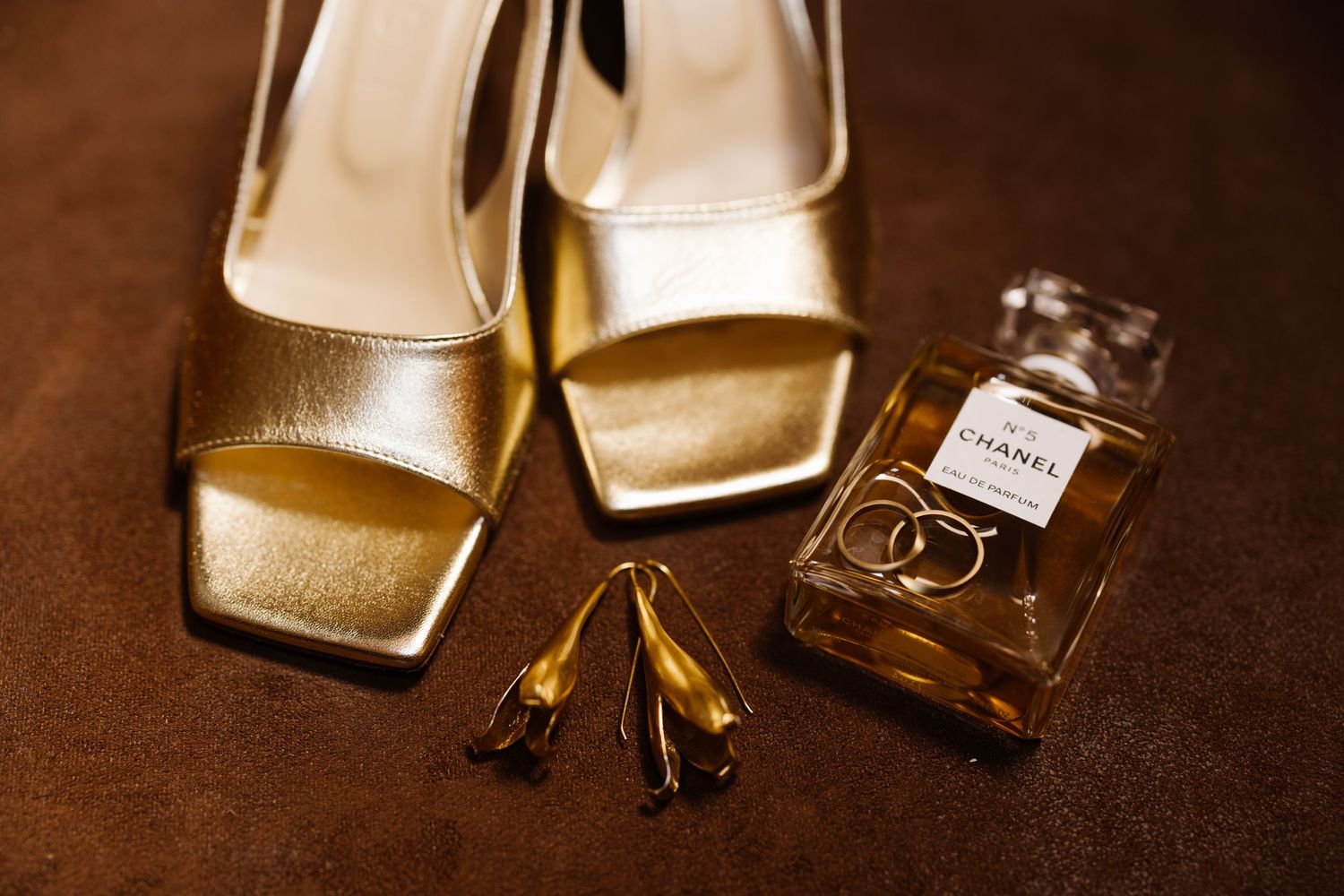 Gold wedding rings and perfume bottles arranged artistically on dark brown leather surface in warm lighting.