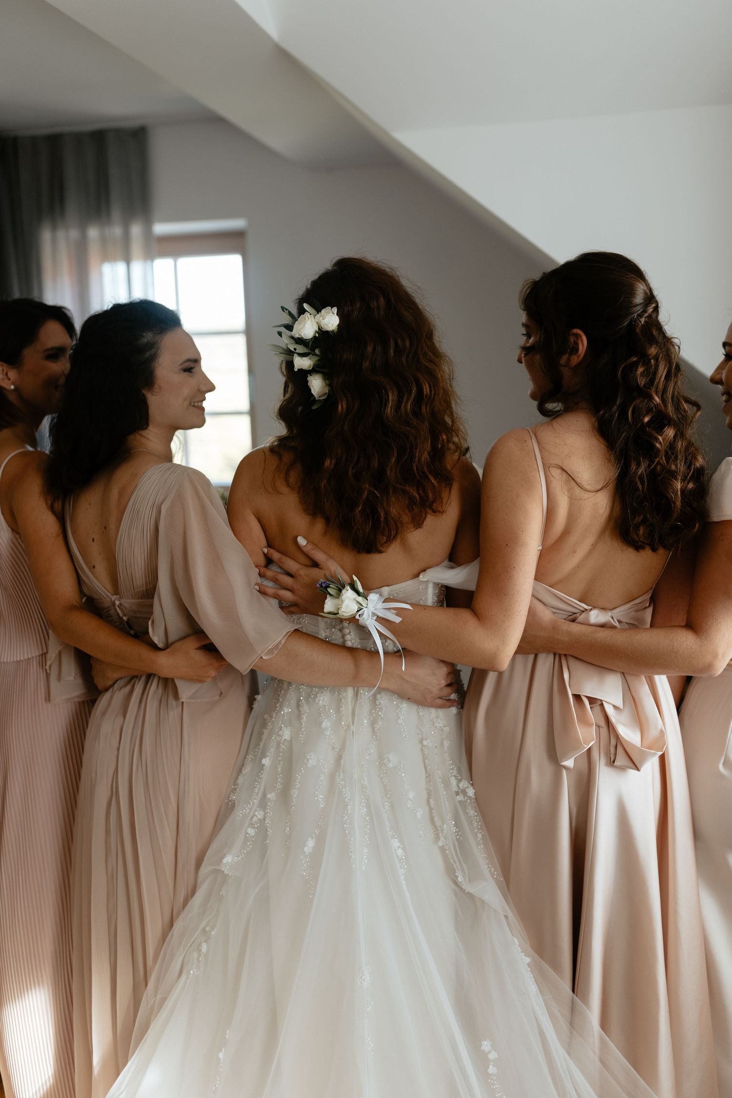 Bridesmaids in blush pink dresses share an intimate group hug in a sunlit room before the wedding ceremony.