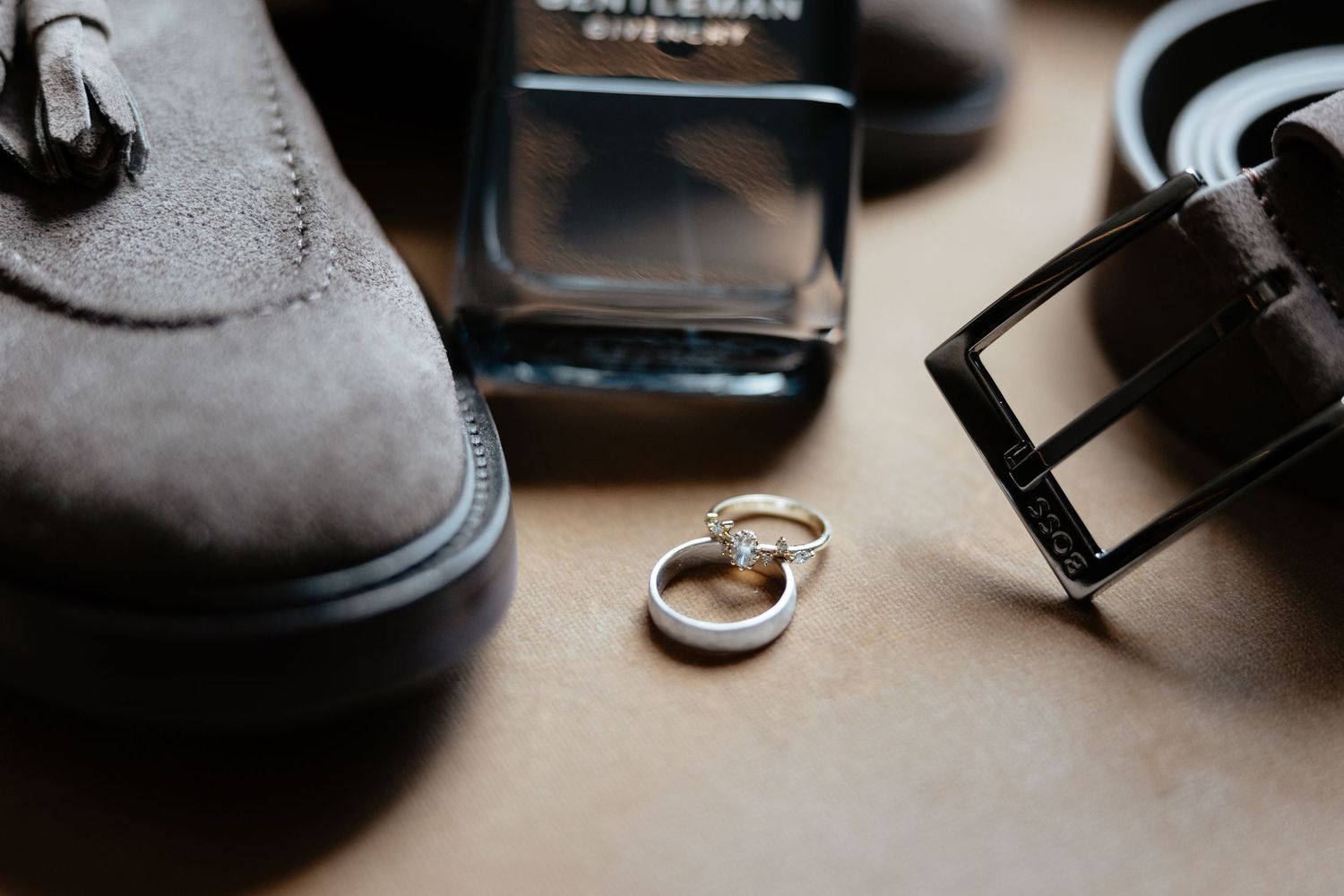 Wedding rings, dress shoes, cologne bottle and leather belt arranged on beige surface in preparation for groom's attire.