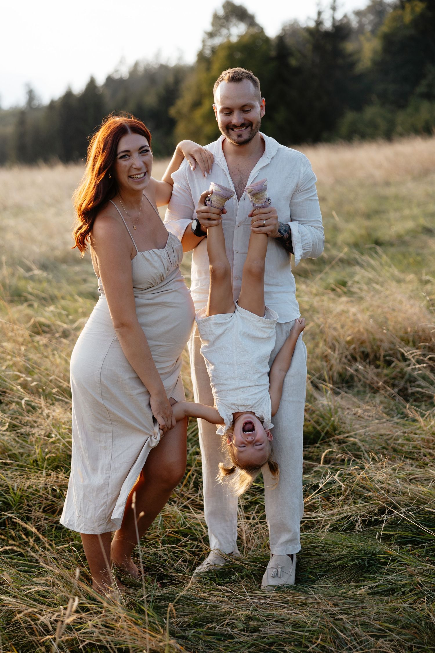A family plays together in a field, playfully holding their child upside down while laughing.