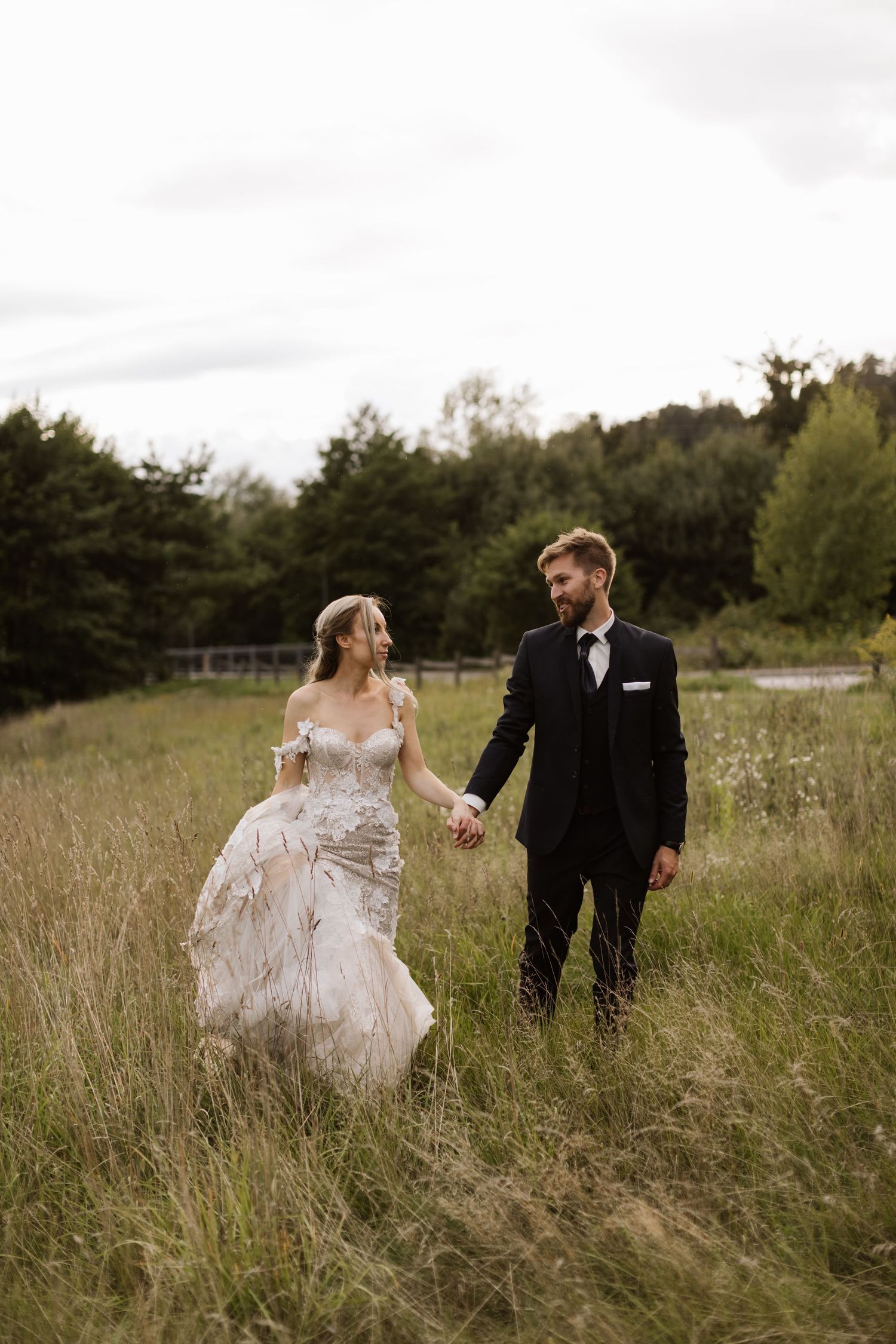 Newlyweds walking hand in hand through a rustic meadow with mountains in the background.