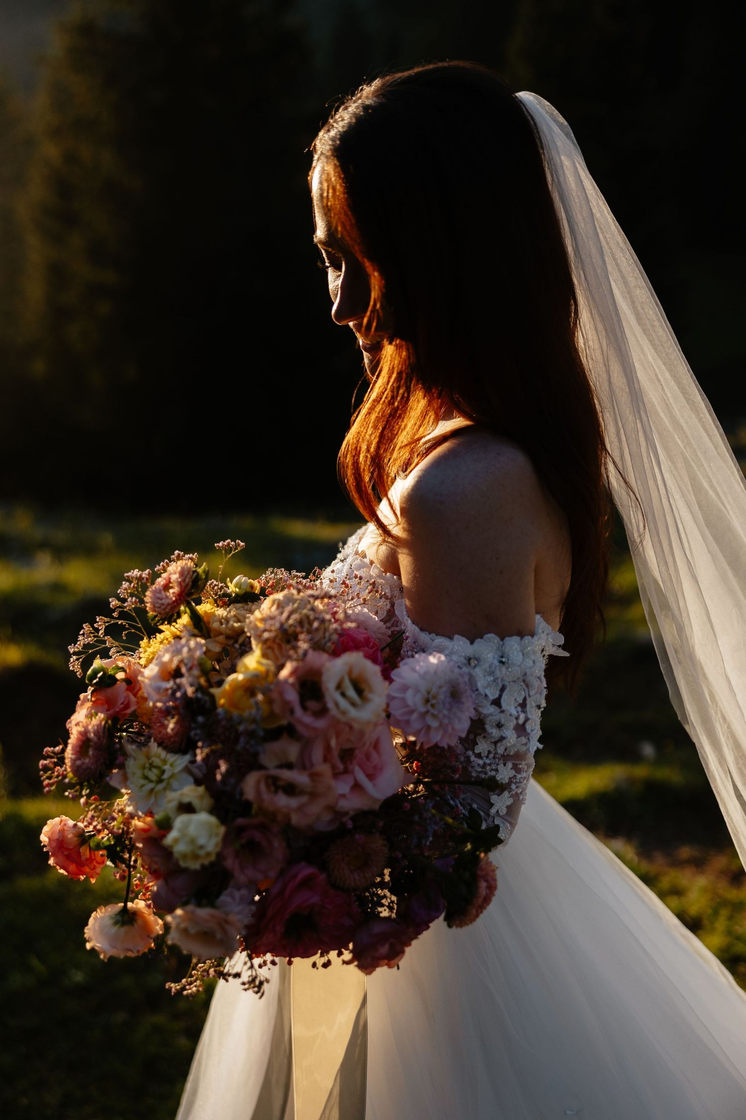 A bride in a white lace gown holds a lush floral bouquet during golden hour.