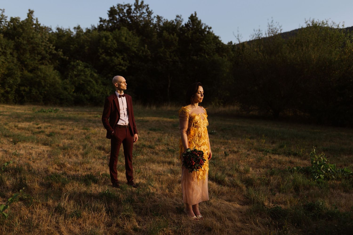 A couple in formal attire stands in a sunlit field during golden hour.