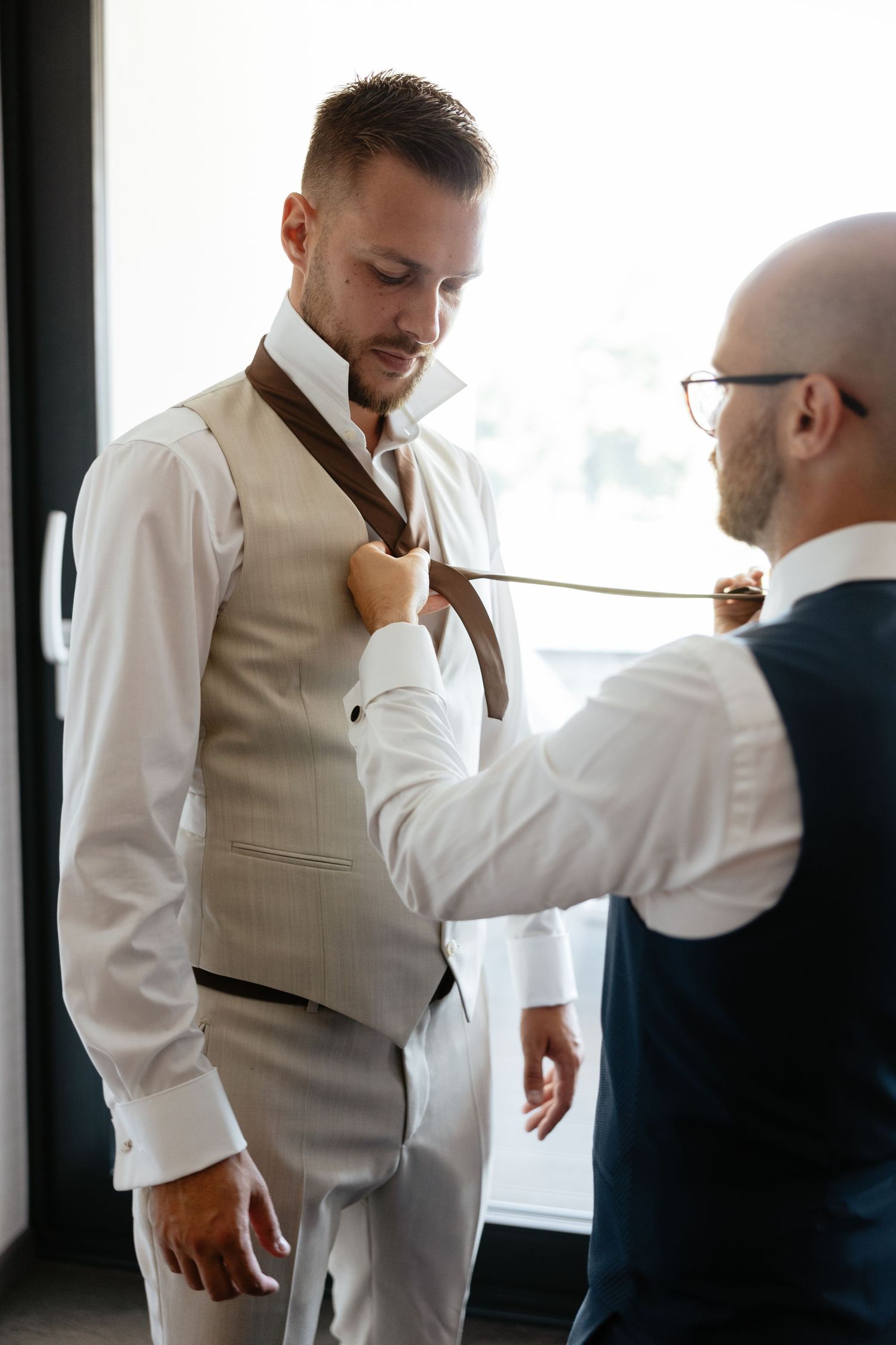 A groomsman helps adjust another's tie during wedding preparations in a sunlit room.