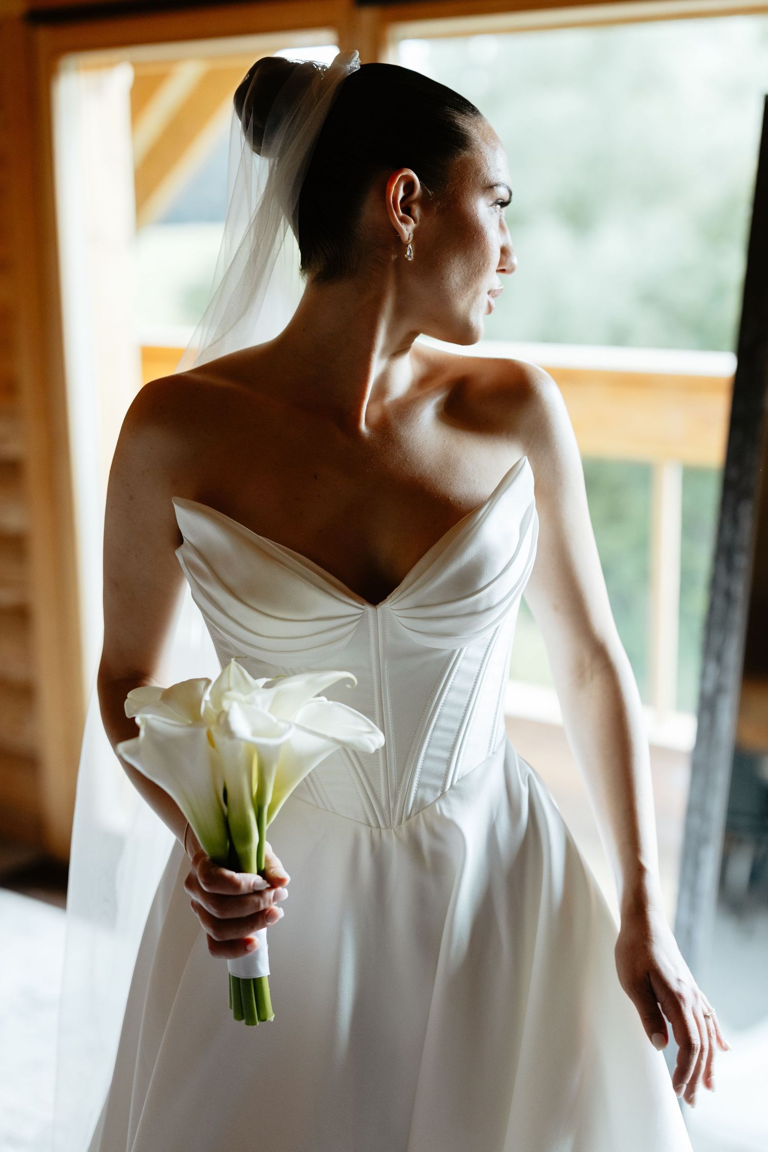 Elegant bride in white wedding dress holds a single calla lily while standing near a window in soft natural light.