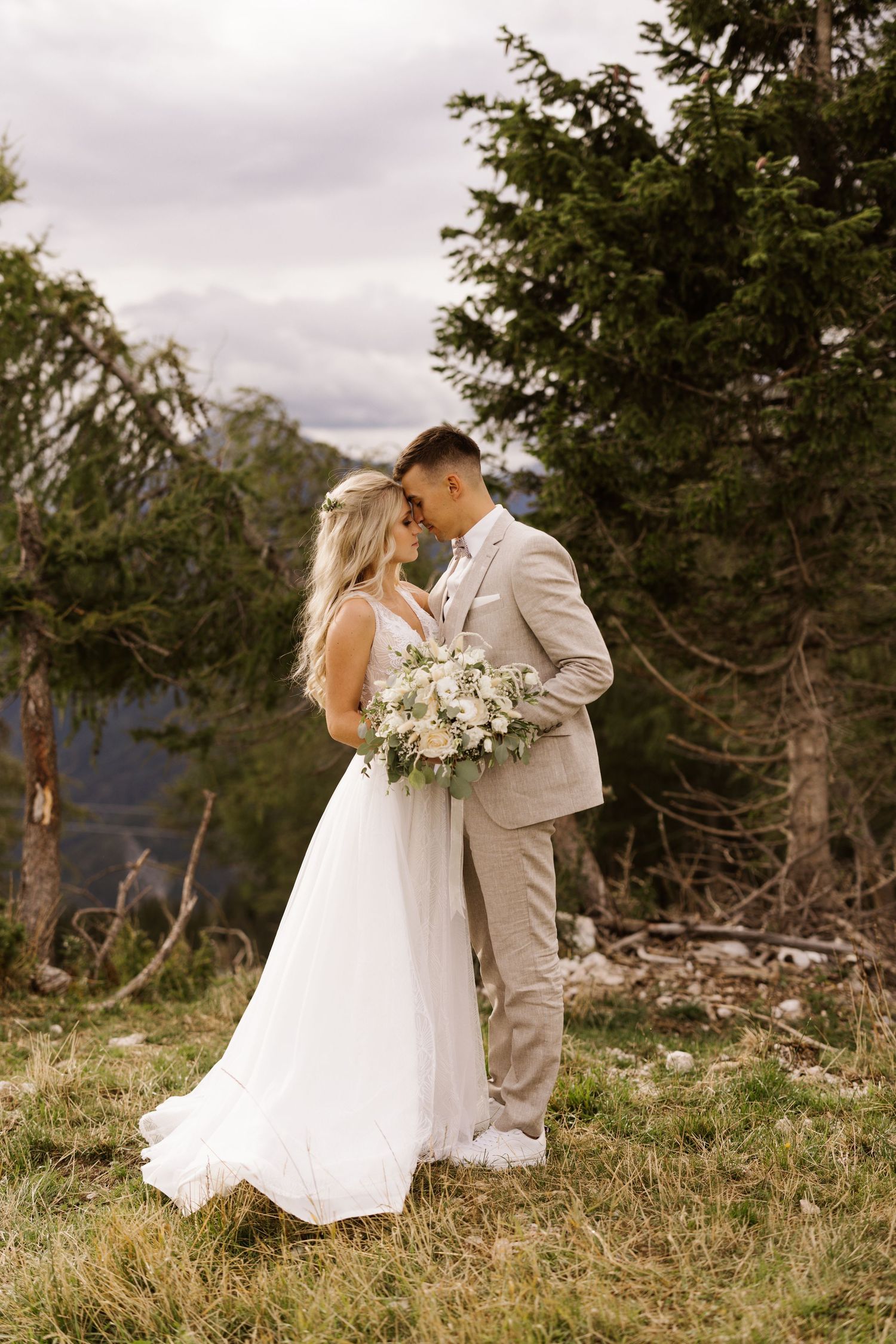 Wedding couple in formal attire embracing on a scenic mountain hillside with evergreen trees.