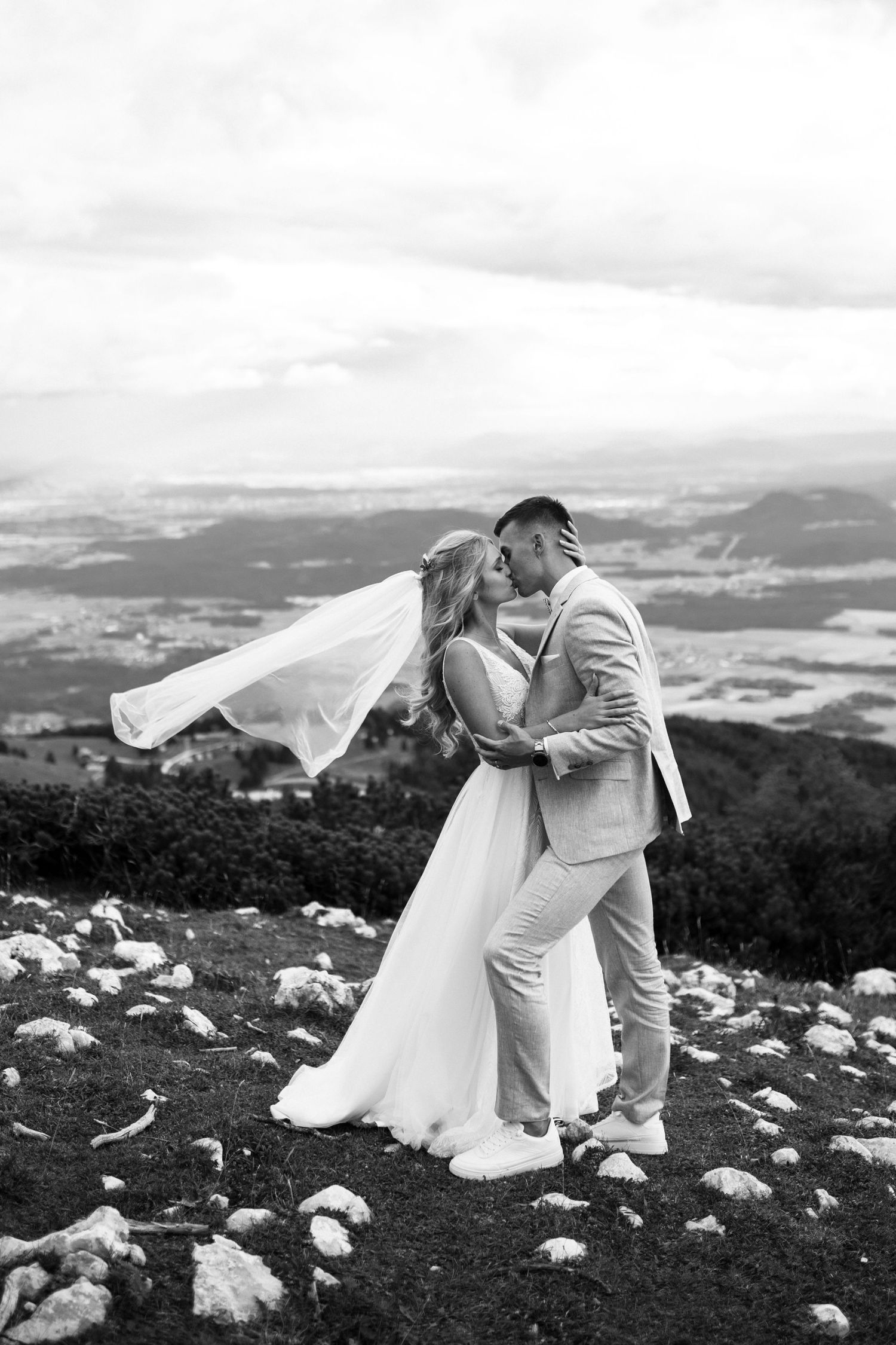 A couple in wedding attire share a romantic moment on a mountain overlook with a dramatic landscape behind them.