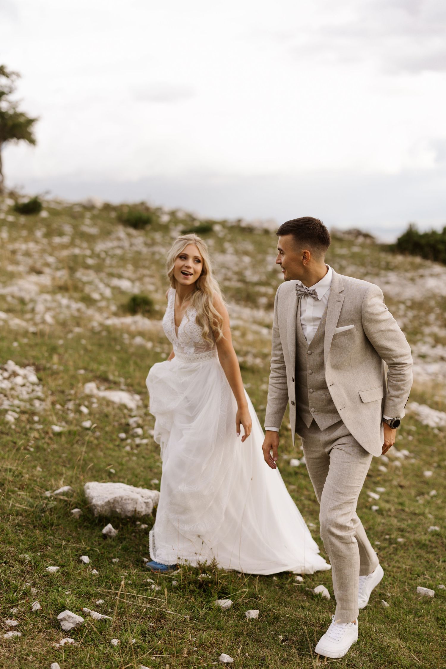 A couple in wedding attire walking together on a grassy hillside with cloudy skies.