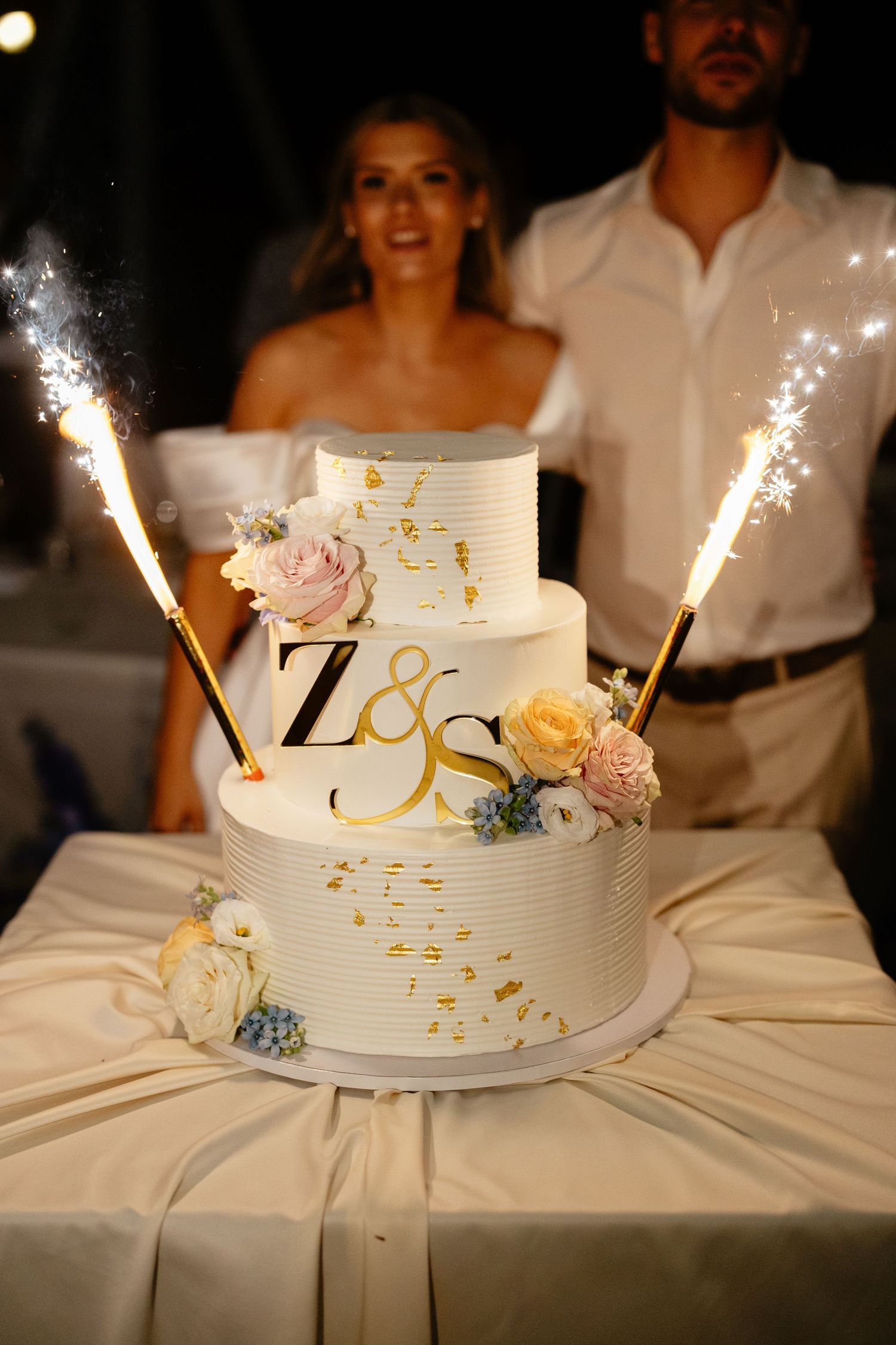A white wedding cake with sparklers and delicate floral decorations displayed in a series of similar shots at night.