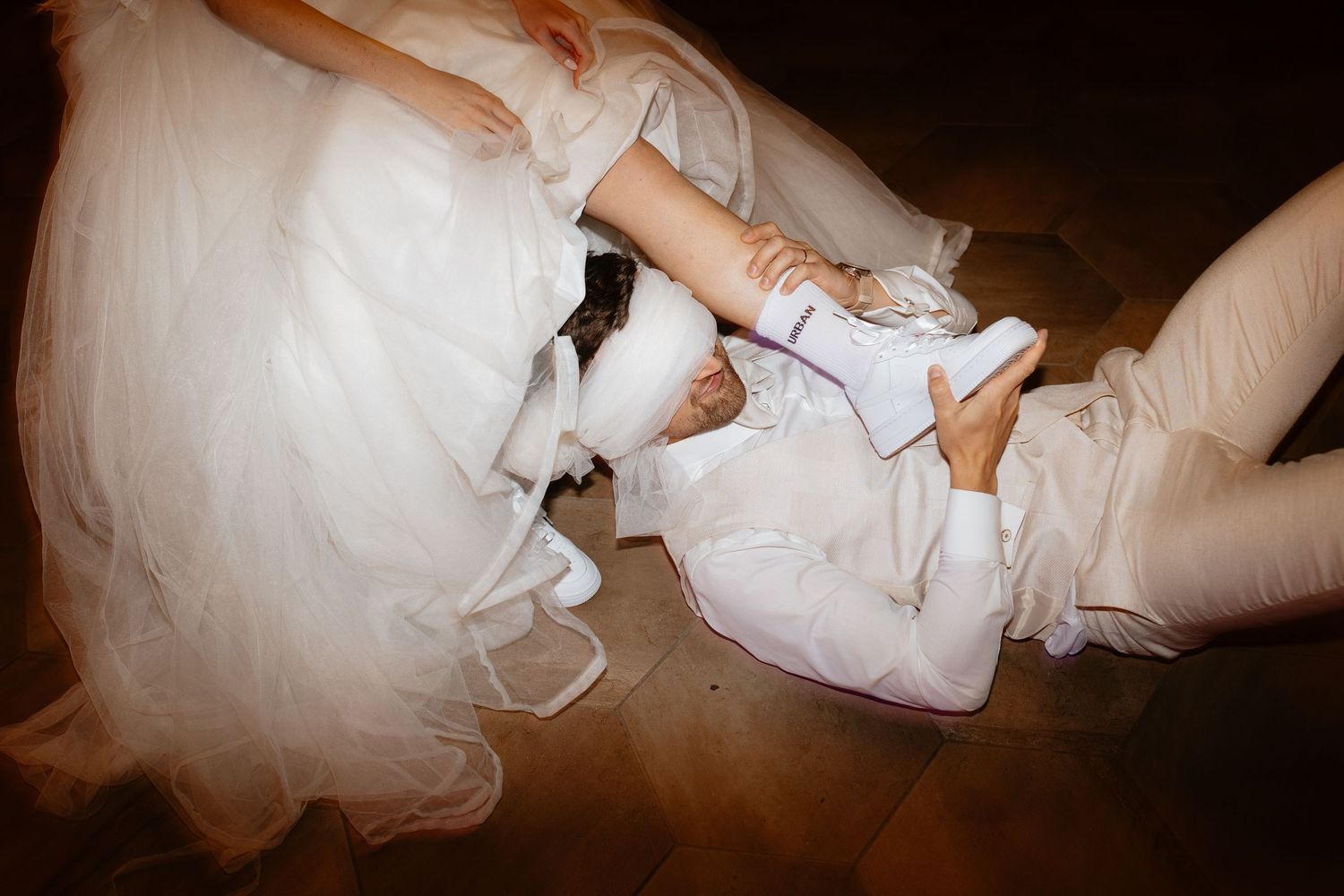 Wedding couple's playful moment showing white dress and formal shoes on a dark floor.