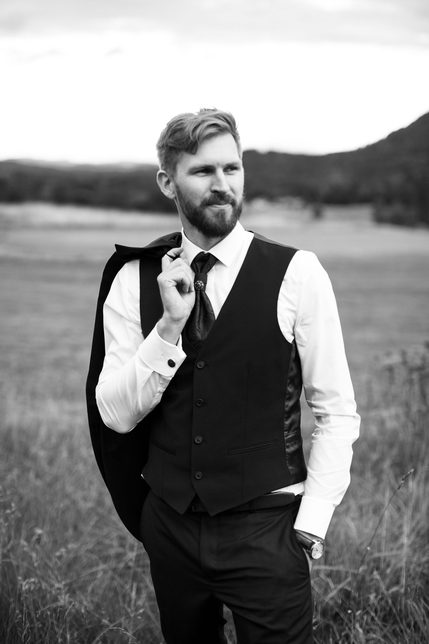Groomsmen in black vests and white shirts pose in a rustic outdoor field setting.