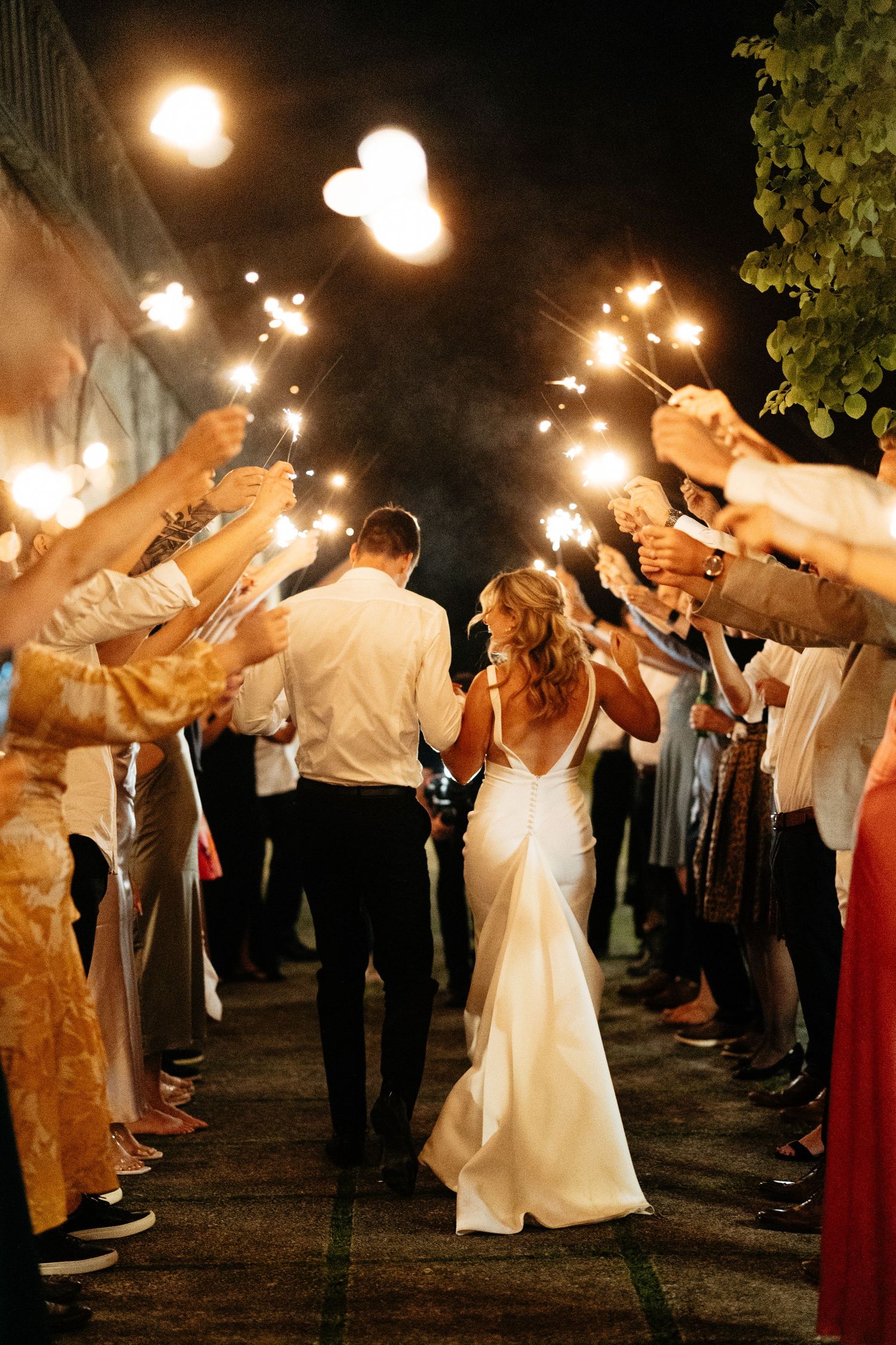 Newlyweds walk through sparkler exit tunnel created by wedding guests at night.