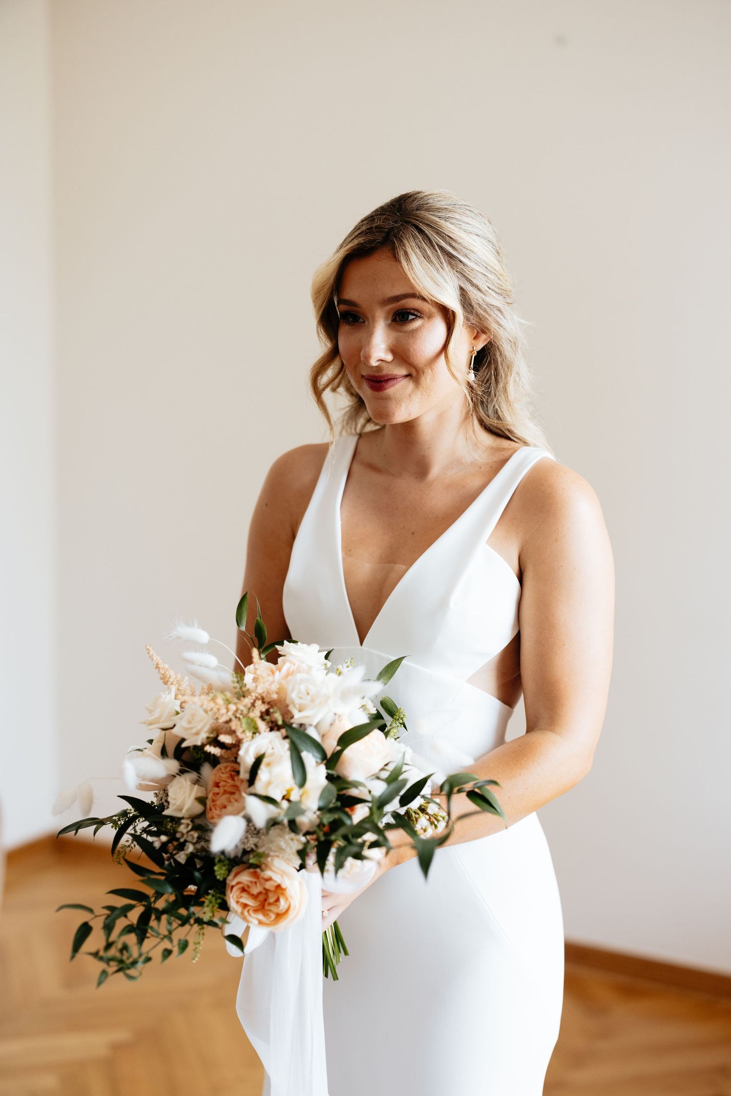 A bride in an elegant white dress holds a romantic bouquet of peach and white flowers.