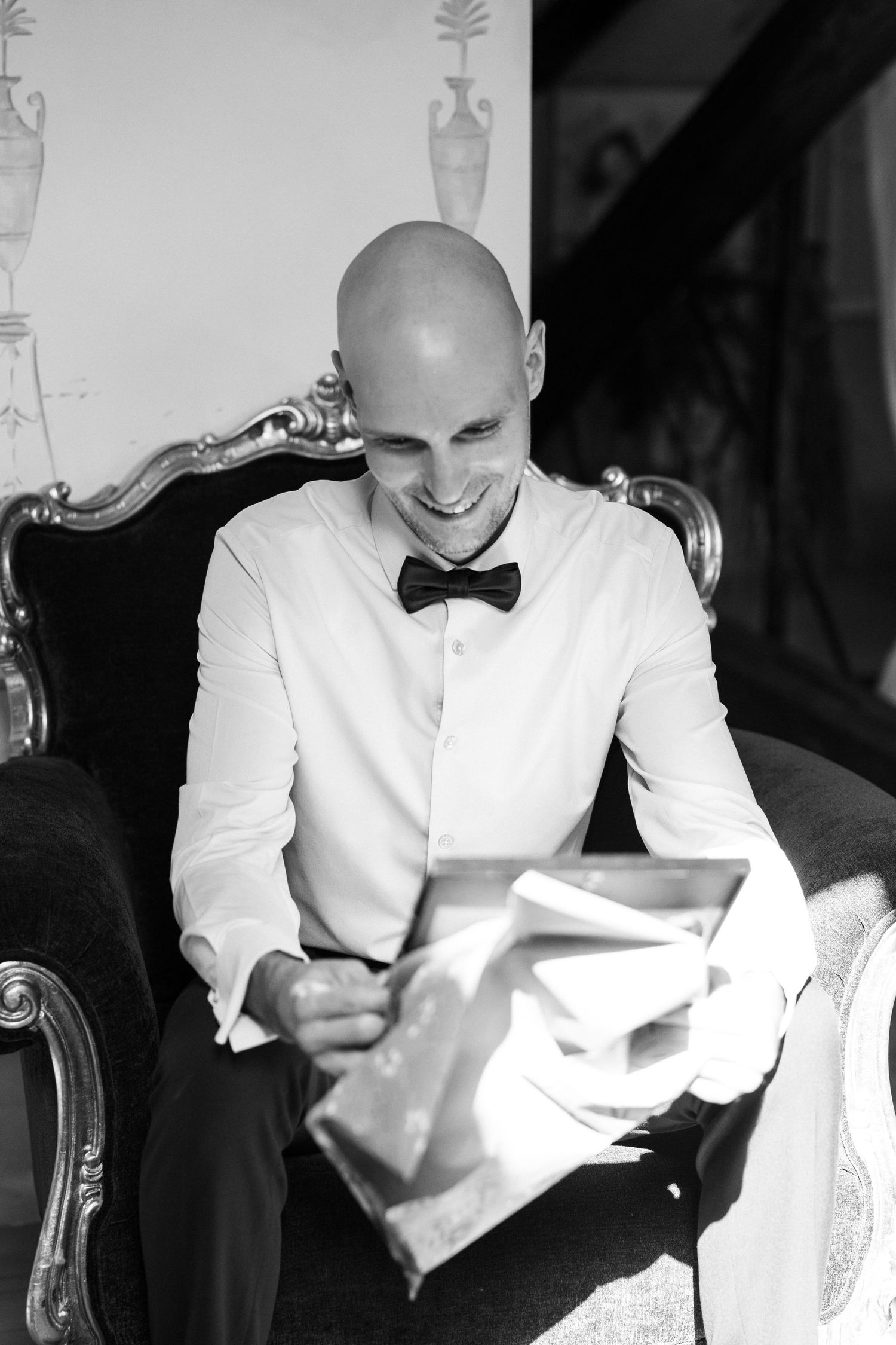 Person in white formal attire and bow tie sitting in an elegant chair during a special event.