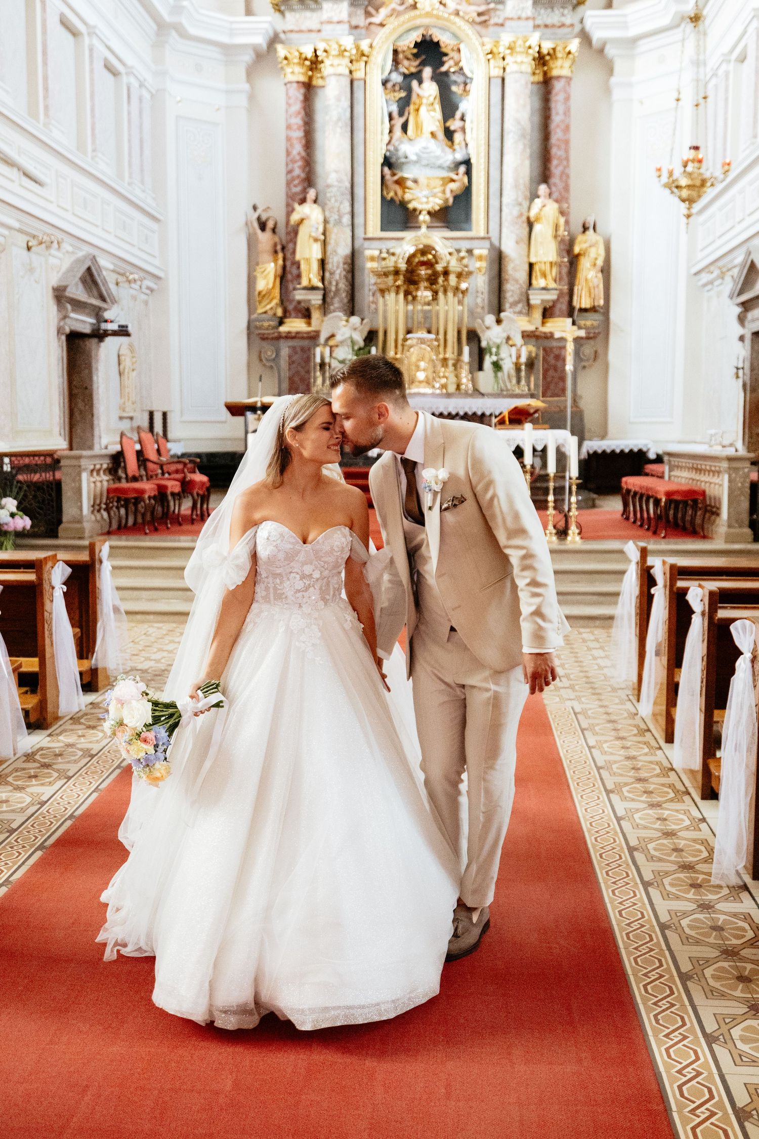 Wedding couple walking down ornate church aisle with white and gold baroque interior decor.