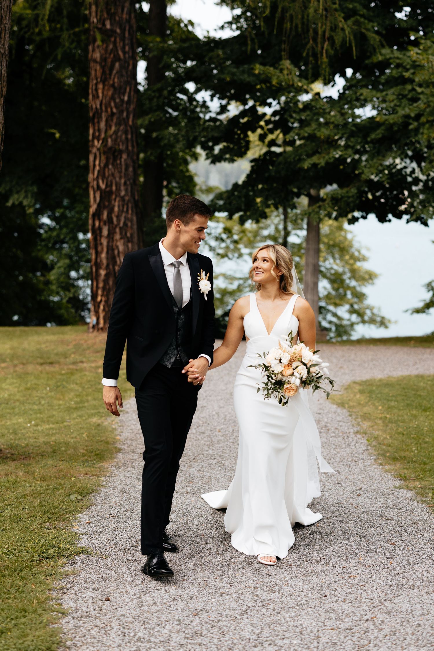 Wedding couple walking together on a gravel path surrounded by tall trees.