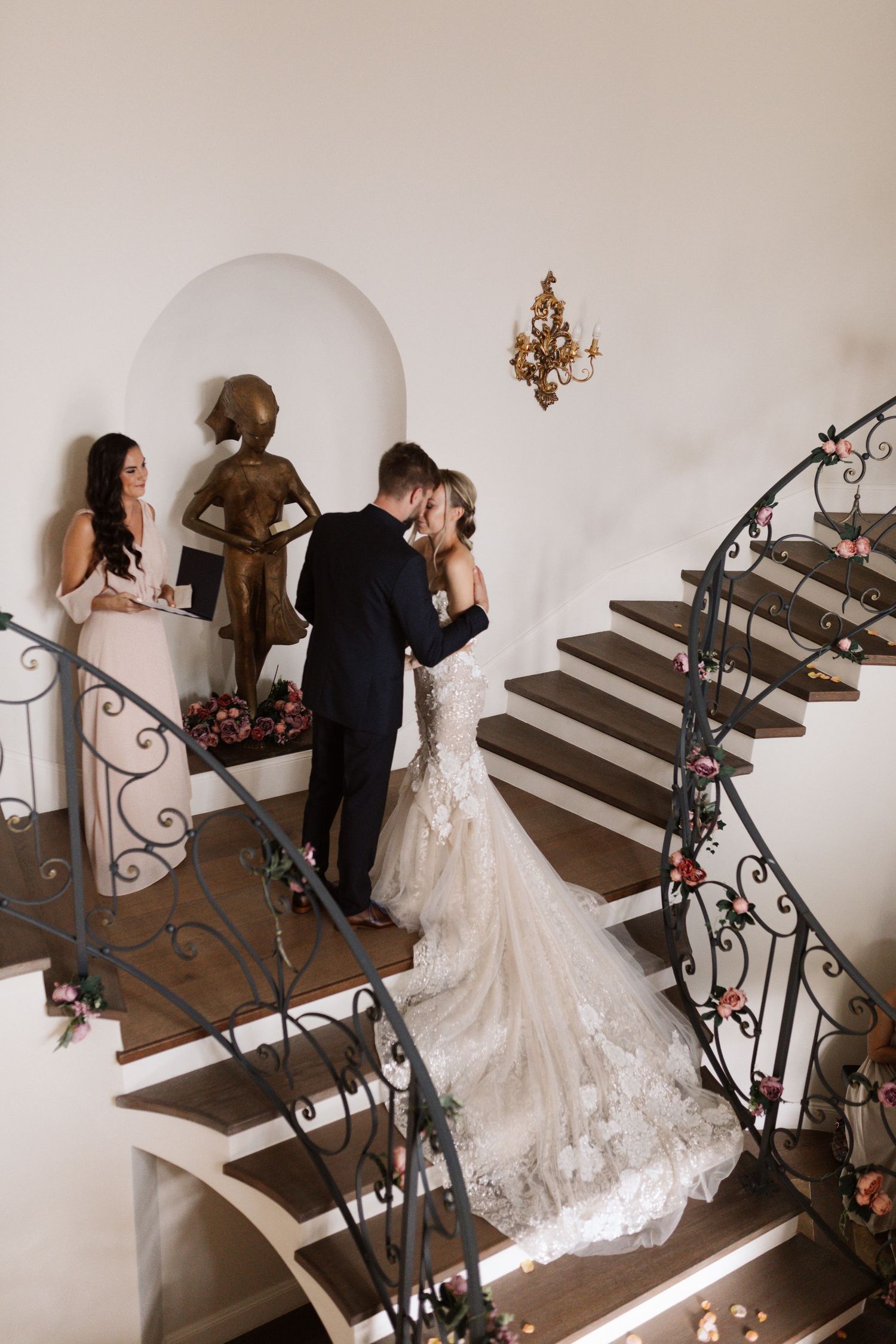 Elegant wedding party descends ornate wooden staircase with wrought iron railings.