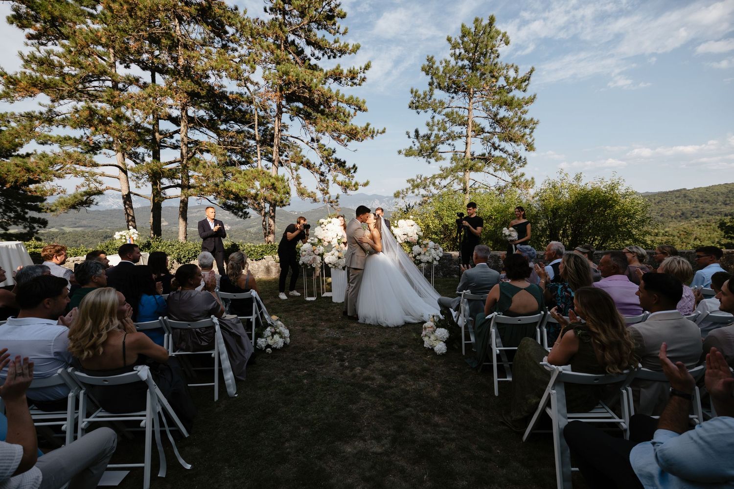 Wide view of outdoor wedding ceremony with guests seated on white chairs among pine trees.