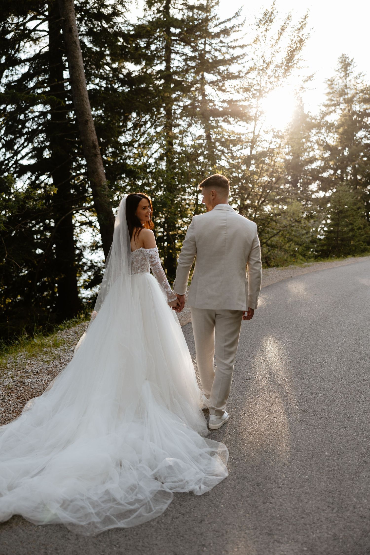 Newlyweds walking hand in hand on a sunlit forest path with a dramatic trailing wedding gown.