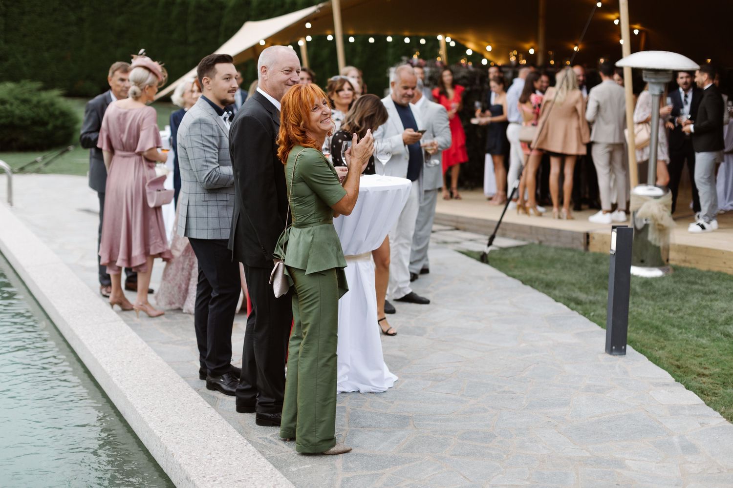 Wedding guests gather around a pool during an elegant outdoor reception with string lights overhead.