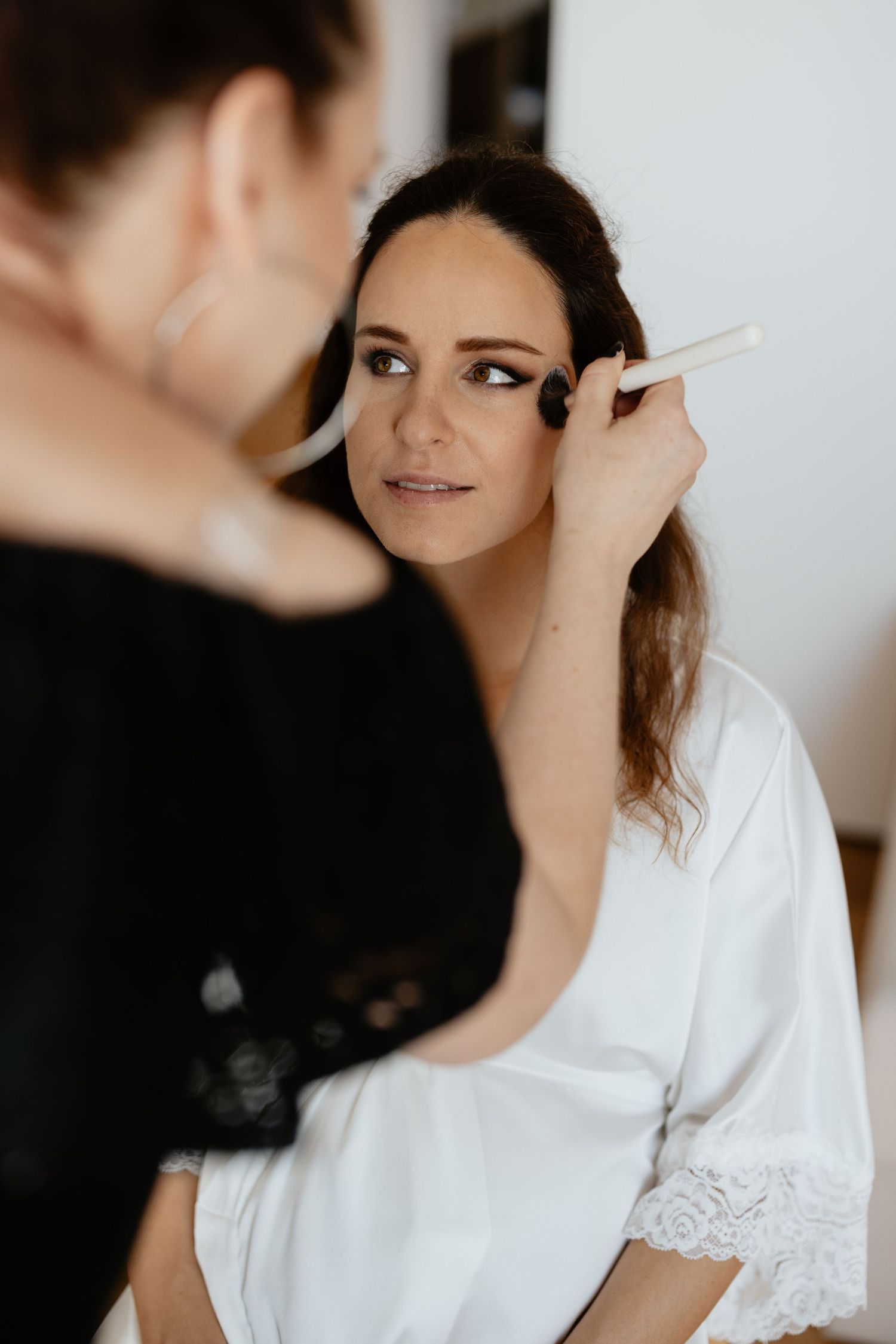 A makeup artist applies eye makeup to a bride wearing a white lace robe during wedding preparations.