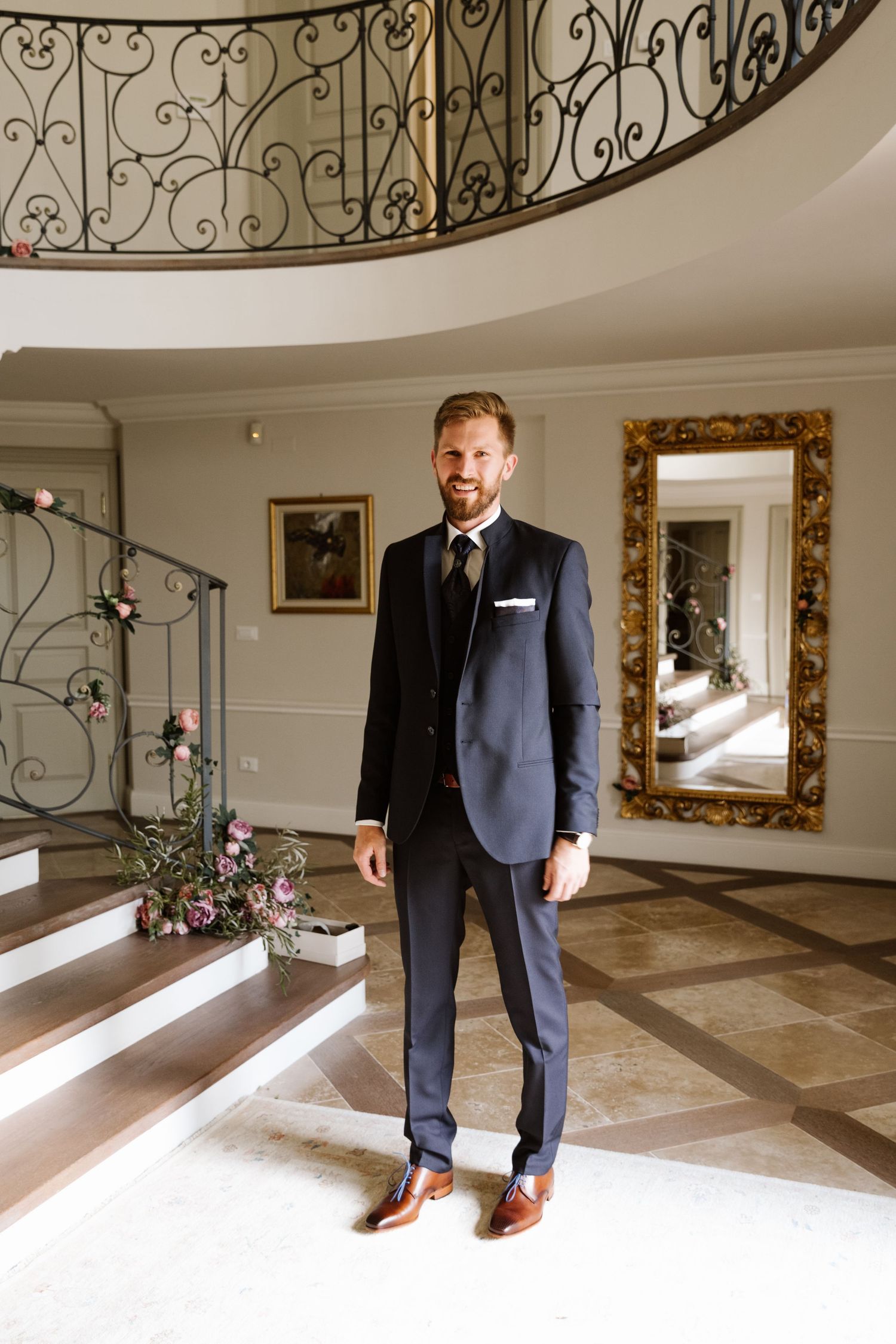 A groom in a classic navy suit stands in an elegant foyer with ornate iron railings and marble floors.