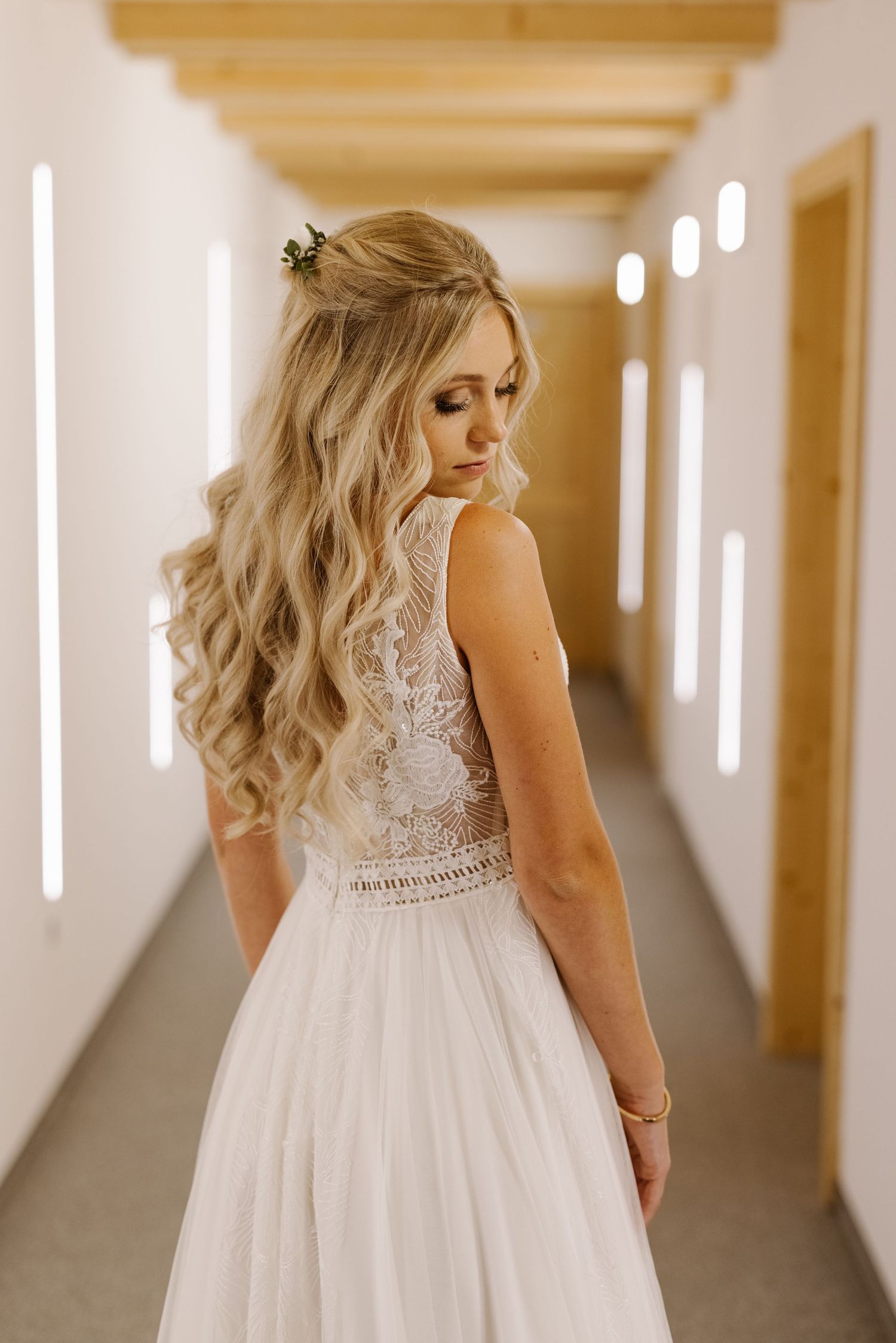 A bride in a white lace wedding dress with flowing blonde hair stands in a sunlit hallway.