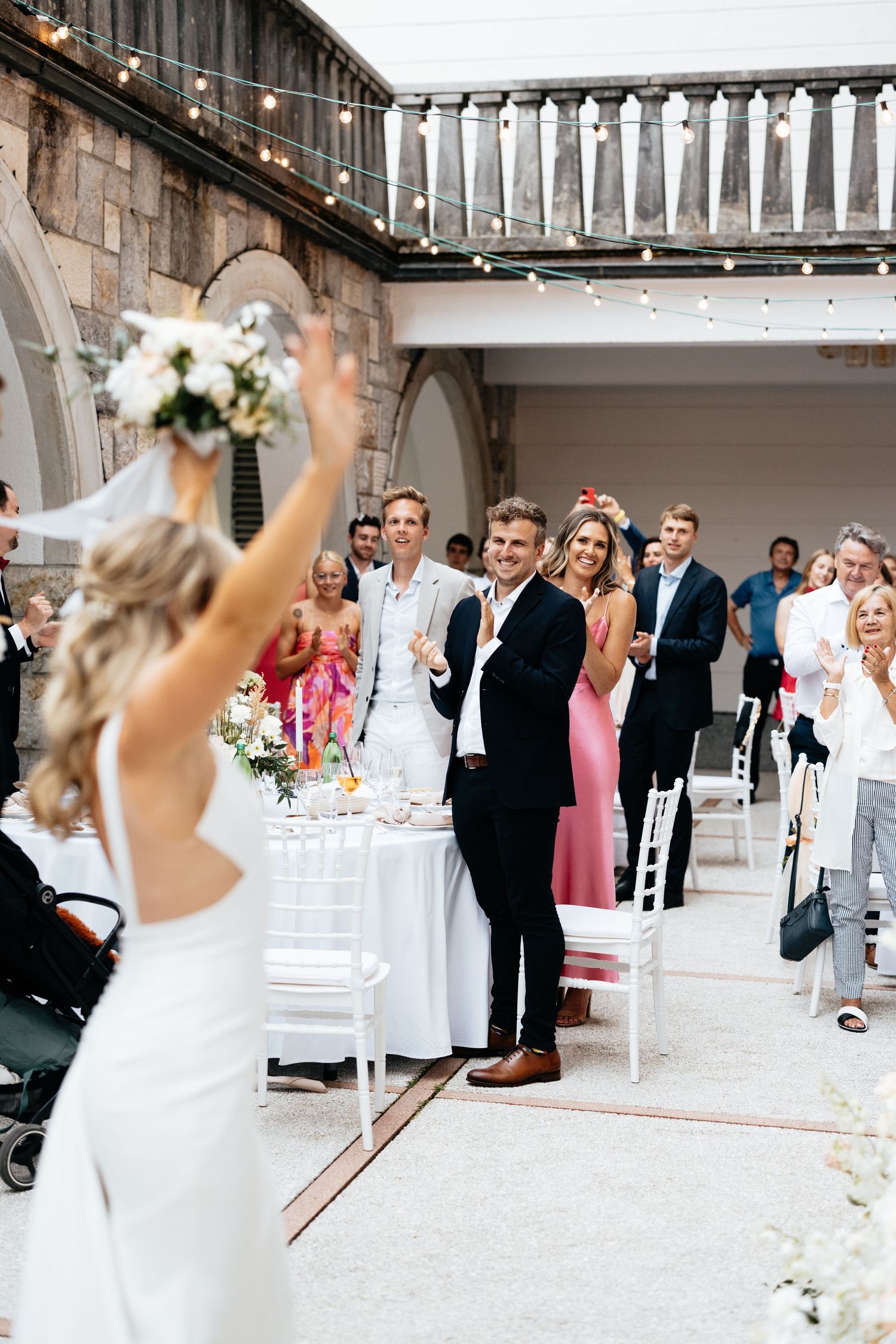 Wedding guests gather in an elegant stone courtyard with white chairs and floral decorations.