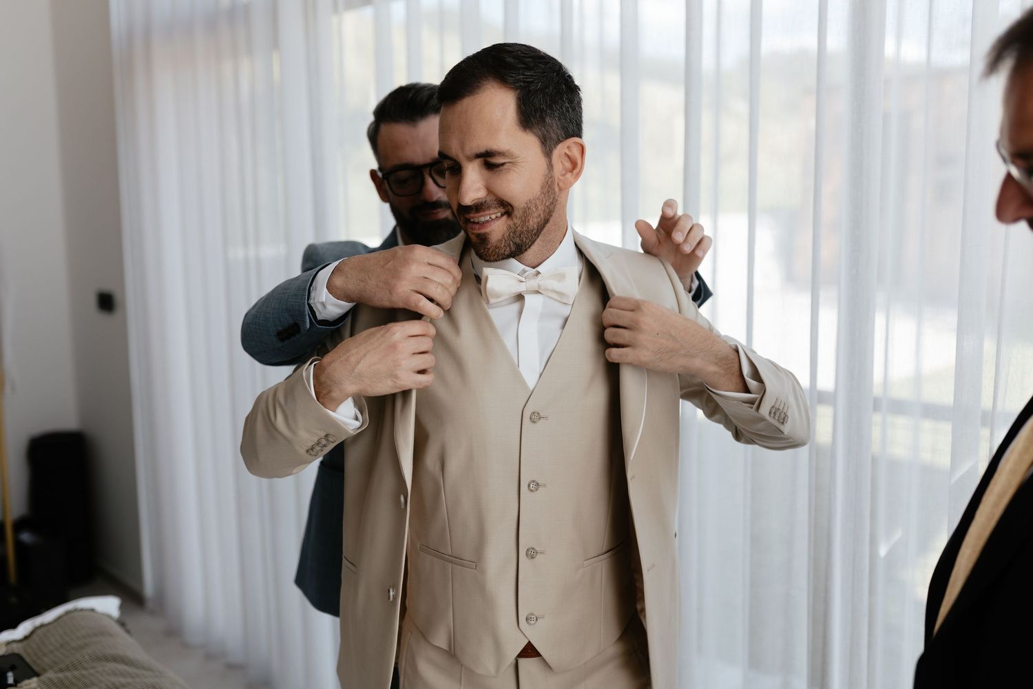 A groom in a beige three-piece suit gets help with final adjustments before the wedding ceremony.