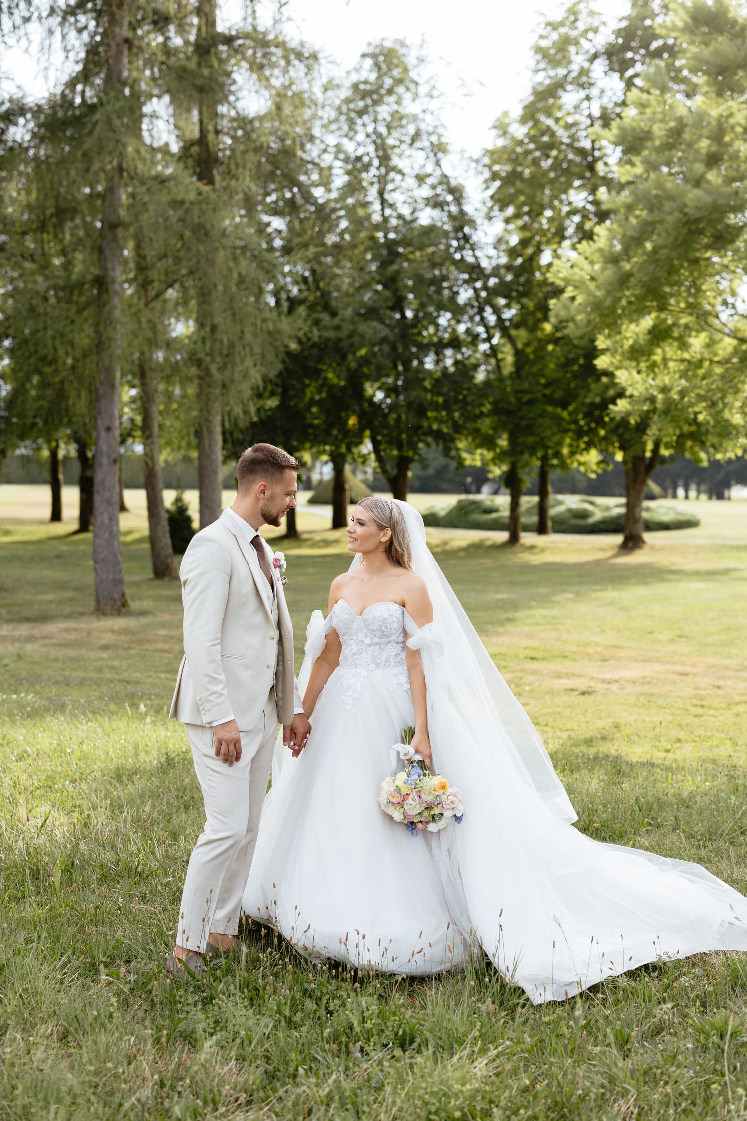 A bride and groom walk hand in hand through a sunlit park setting with lush green trees in the background.