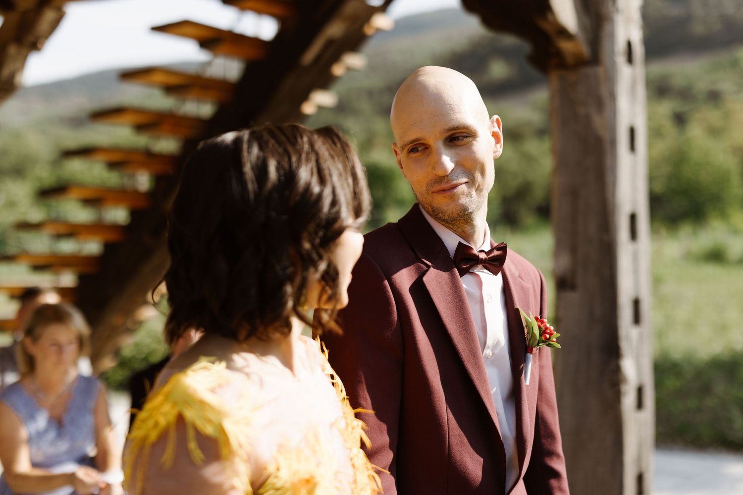 A couple shares an intimate moment under a wooden pergola during an outdoor wedding ceremony.