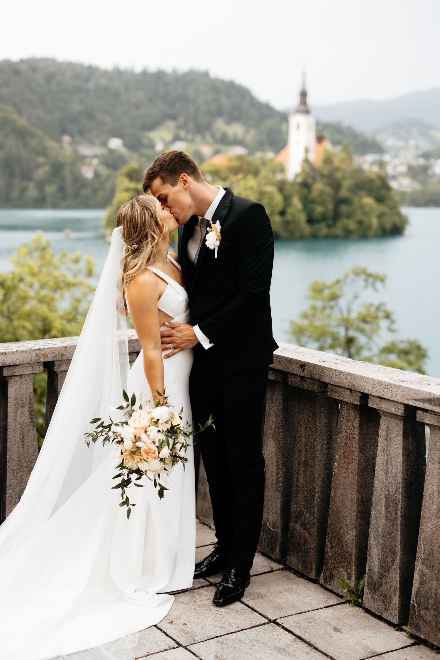 Romantic wedding moment on a balcony overlooking a lake with a church spire in the background.