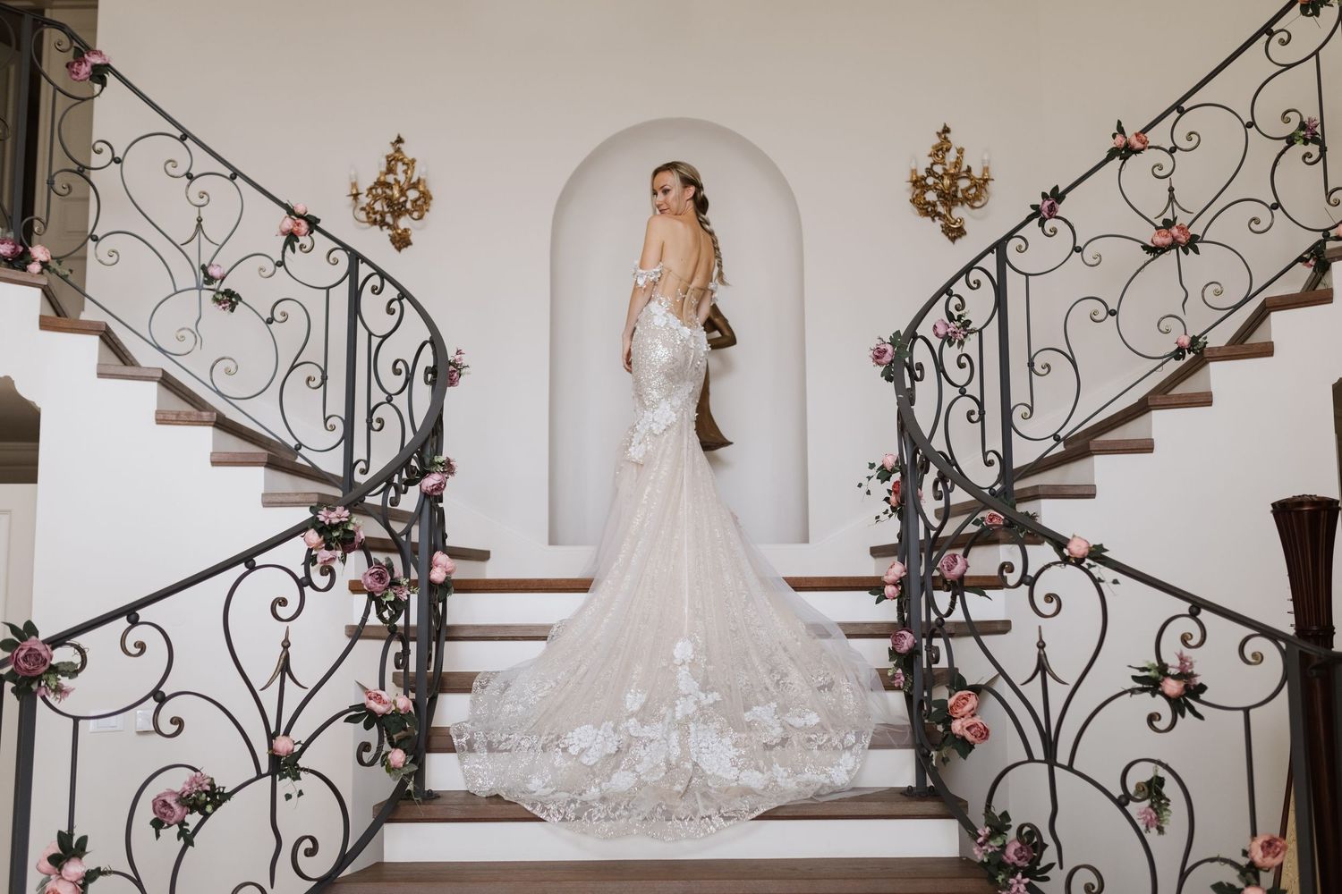 A bride in a flowing white gown poses on an elegant staircase with ornate wrought iron railings and floral decorations.