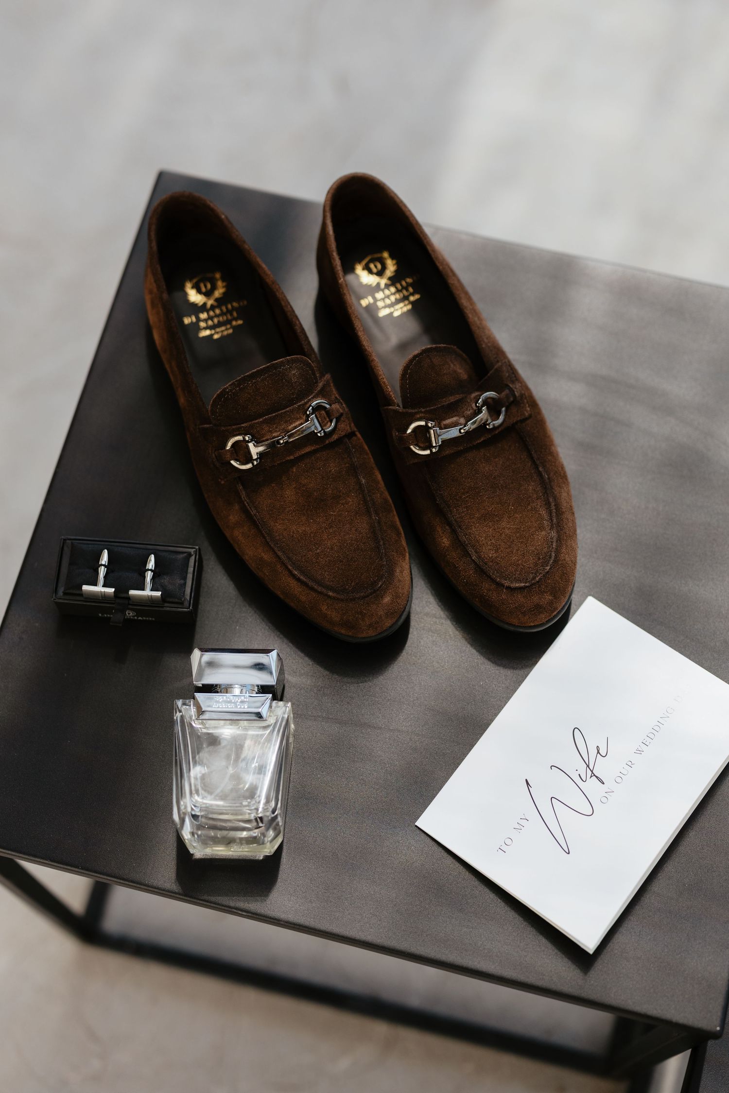 Brown suede loafers displayed next to a glass perfume bottle on a dark wooden surface.