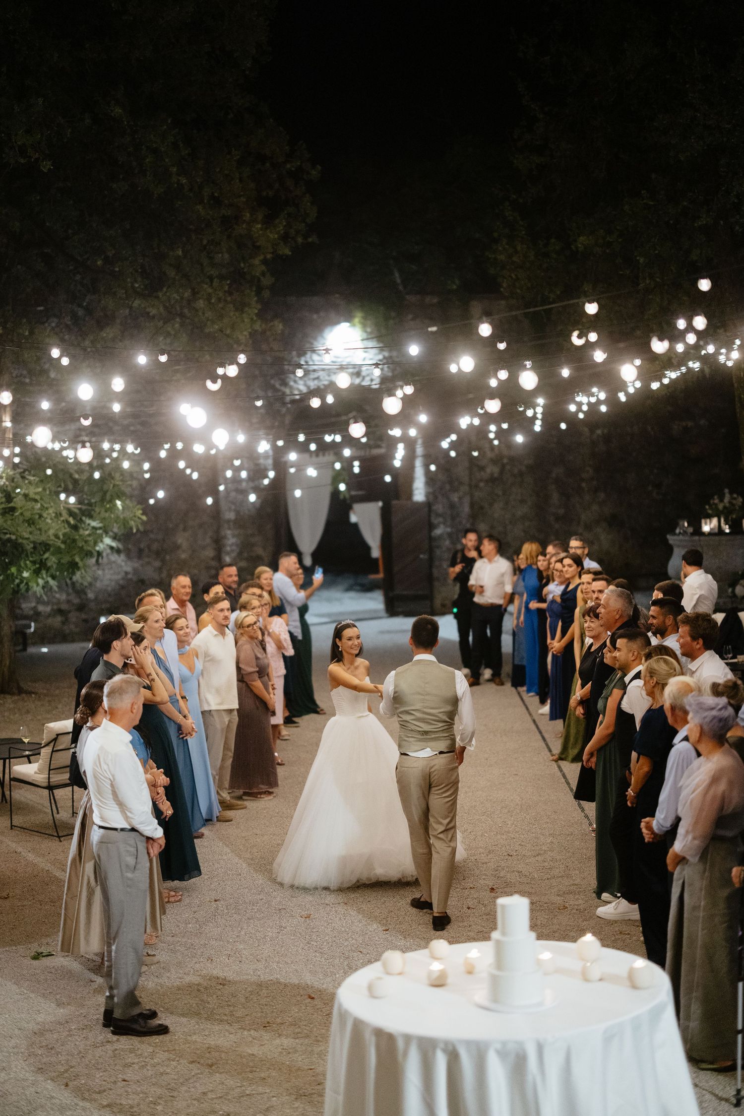 Wedding guests gather around a cake table under twinkling lights at an outdoor reception.