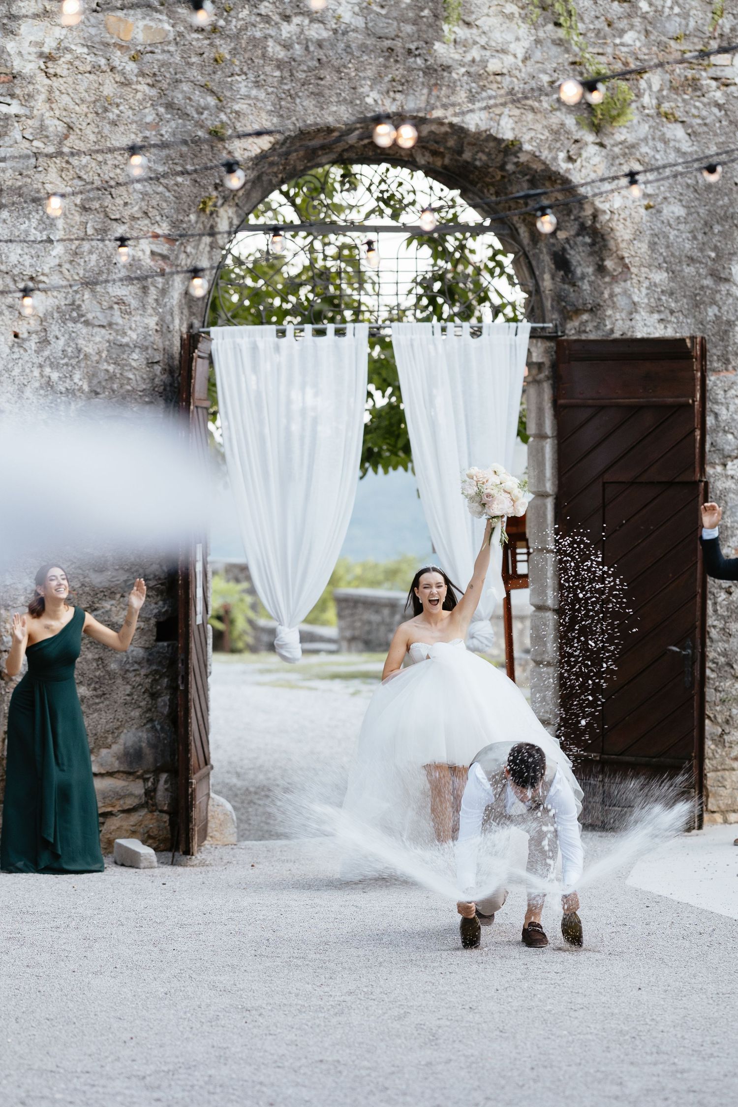 Wedding party celebrates with confetti toss in front of rustic stone wall with string lights.