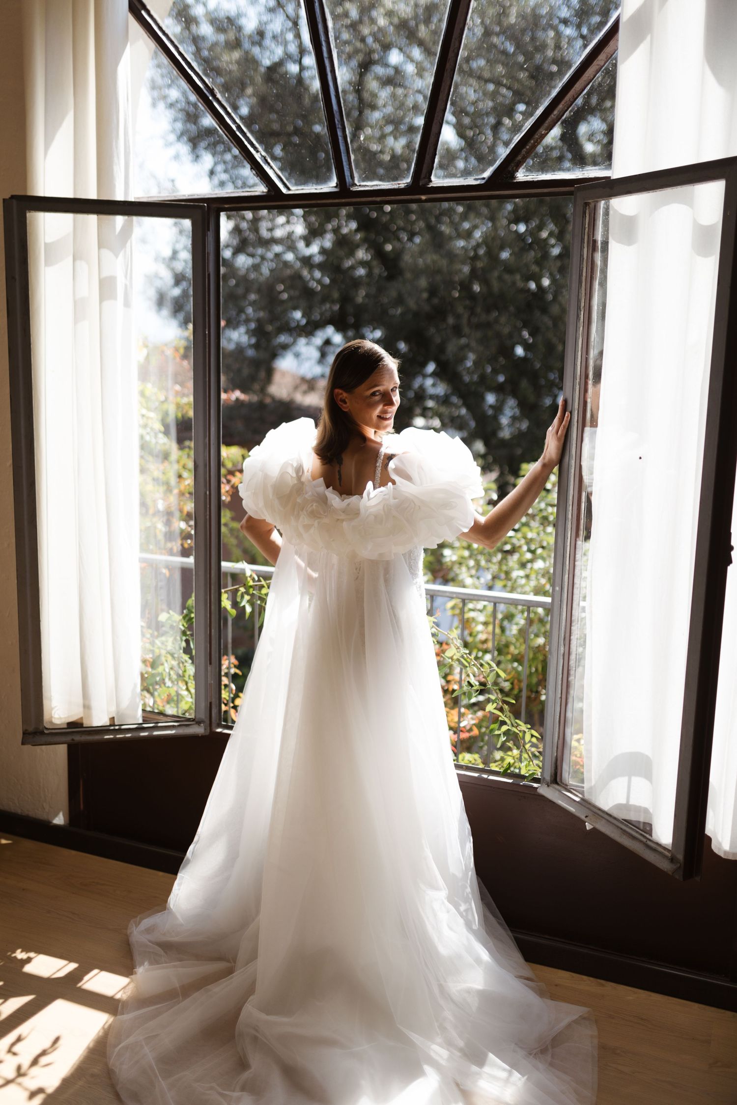 Elegant white wedding gown with dramatic puff sleeves photographed in sunlit window alcove with garden views.