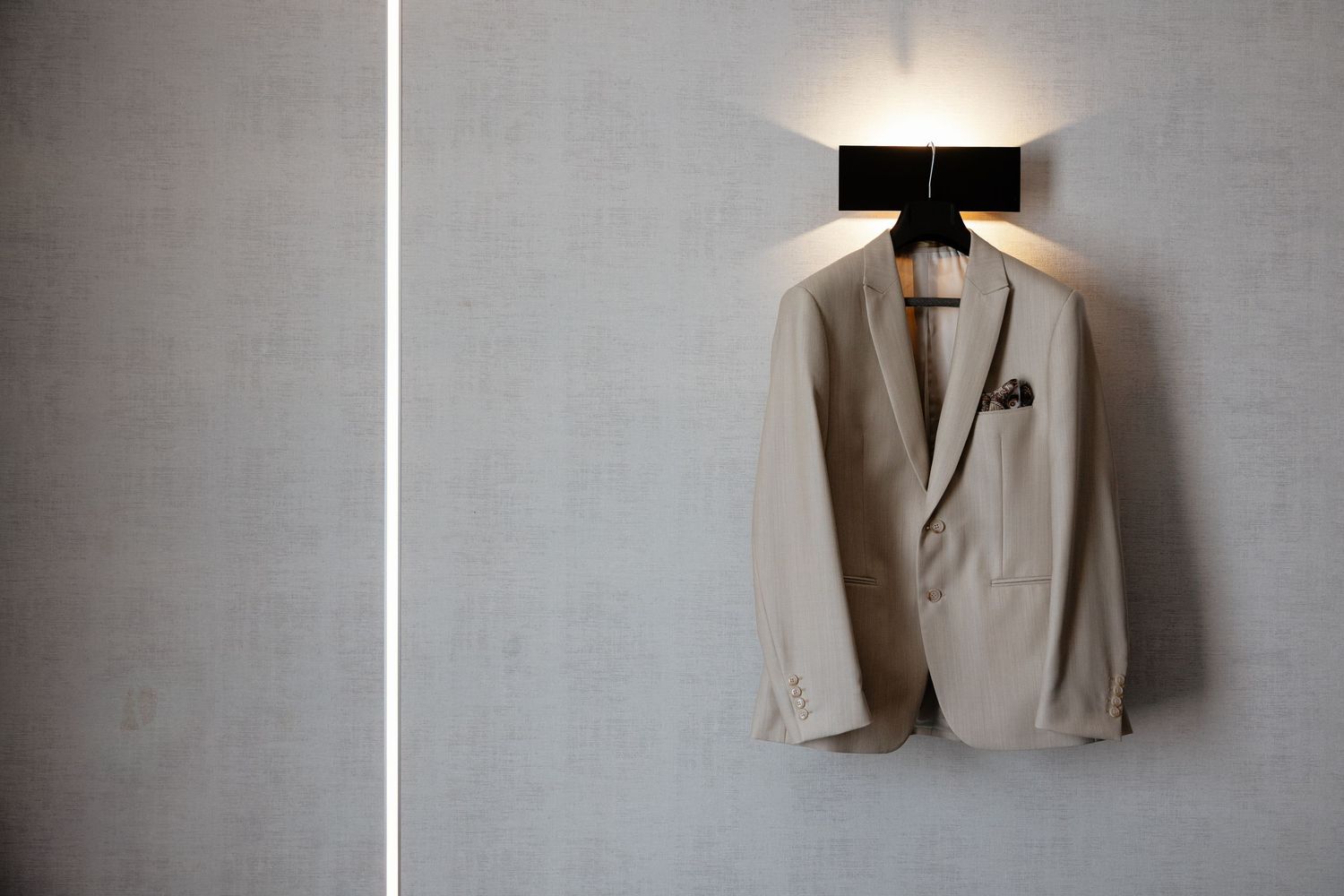 A beige suit jacket hangs elegantly against a gray wall, showcasing classic tailoring and a lapel pin.