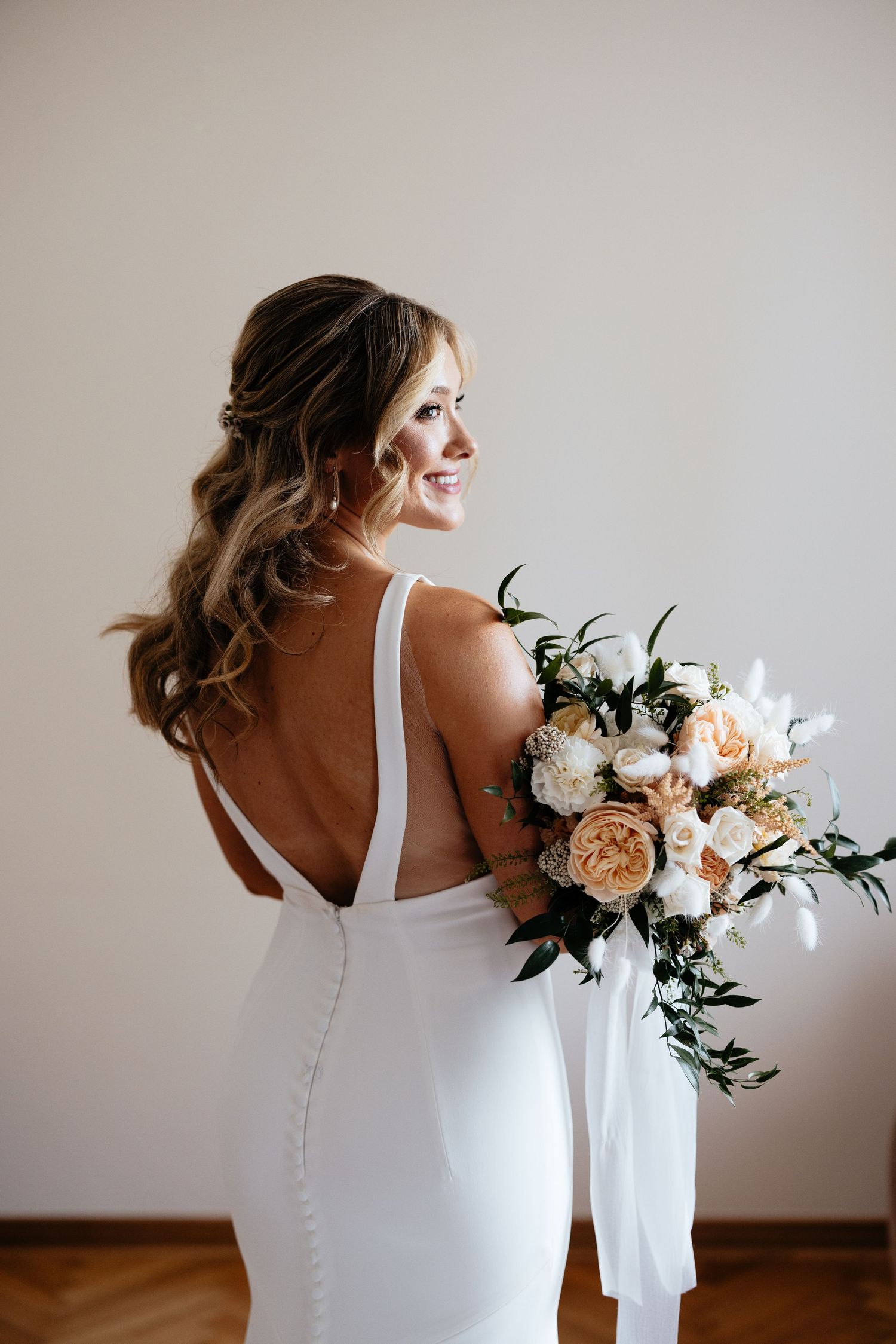 Elegant bride in white dress holding a peach and white floral bouquet against neutral wall.