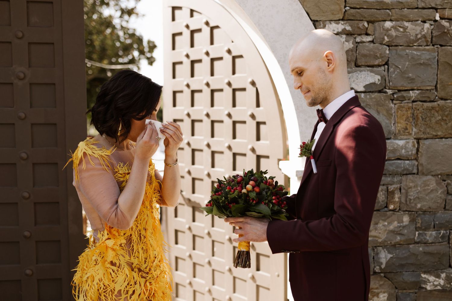 A couple in formal attire dances together in an outdoor setting with stone walls and decorative lattice.