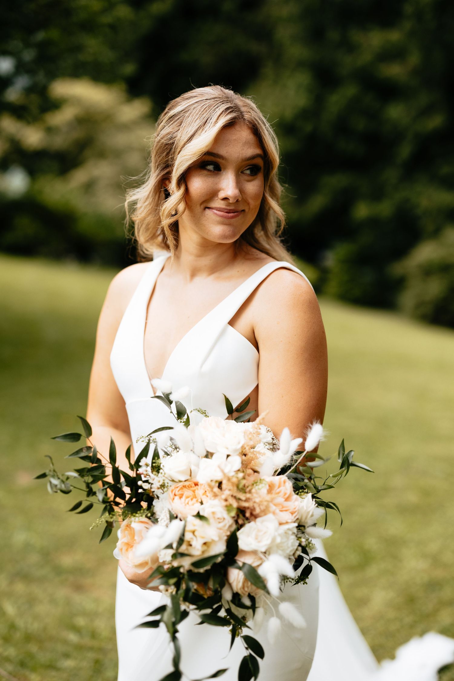 Bride in elegant white dress holds a peach and white floral bouquet with greenery outdoors.