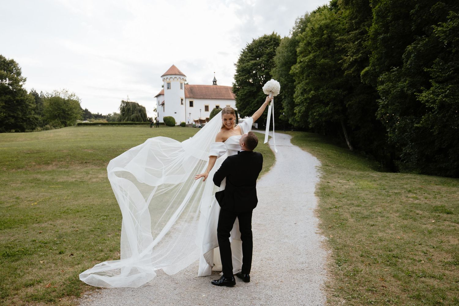 A couple walks along a gravel path with a white church steeple visible in the background on their wedding day.